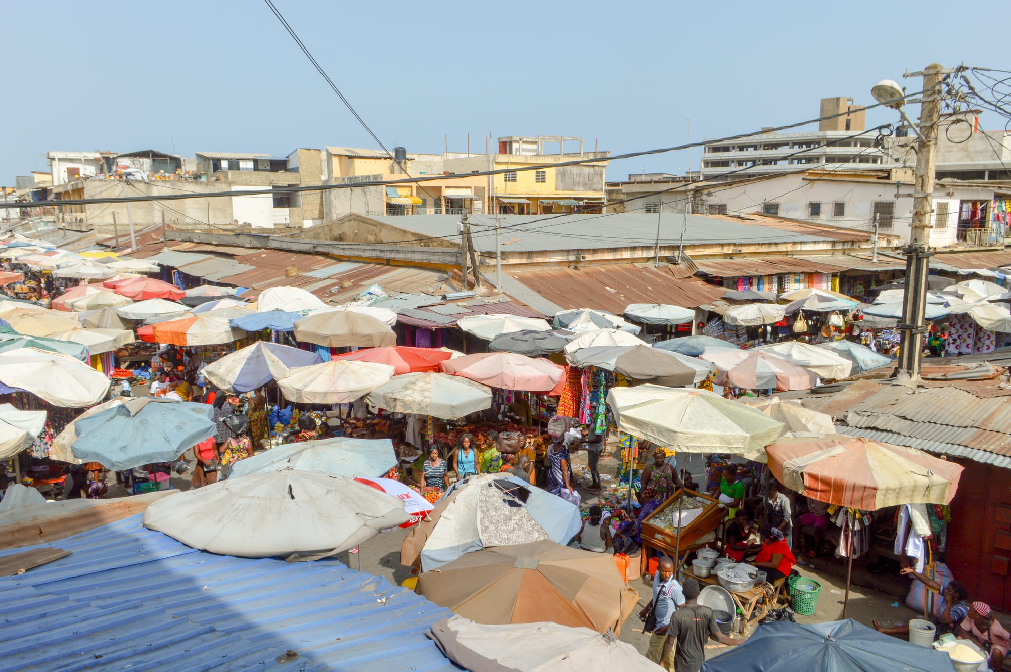 An elevated view over a dense canopy of market umbrellas and corrugated rooftops in Lomé, with shoppers visible between the stalls