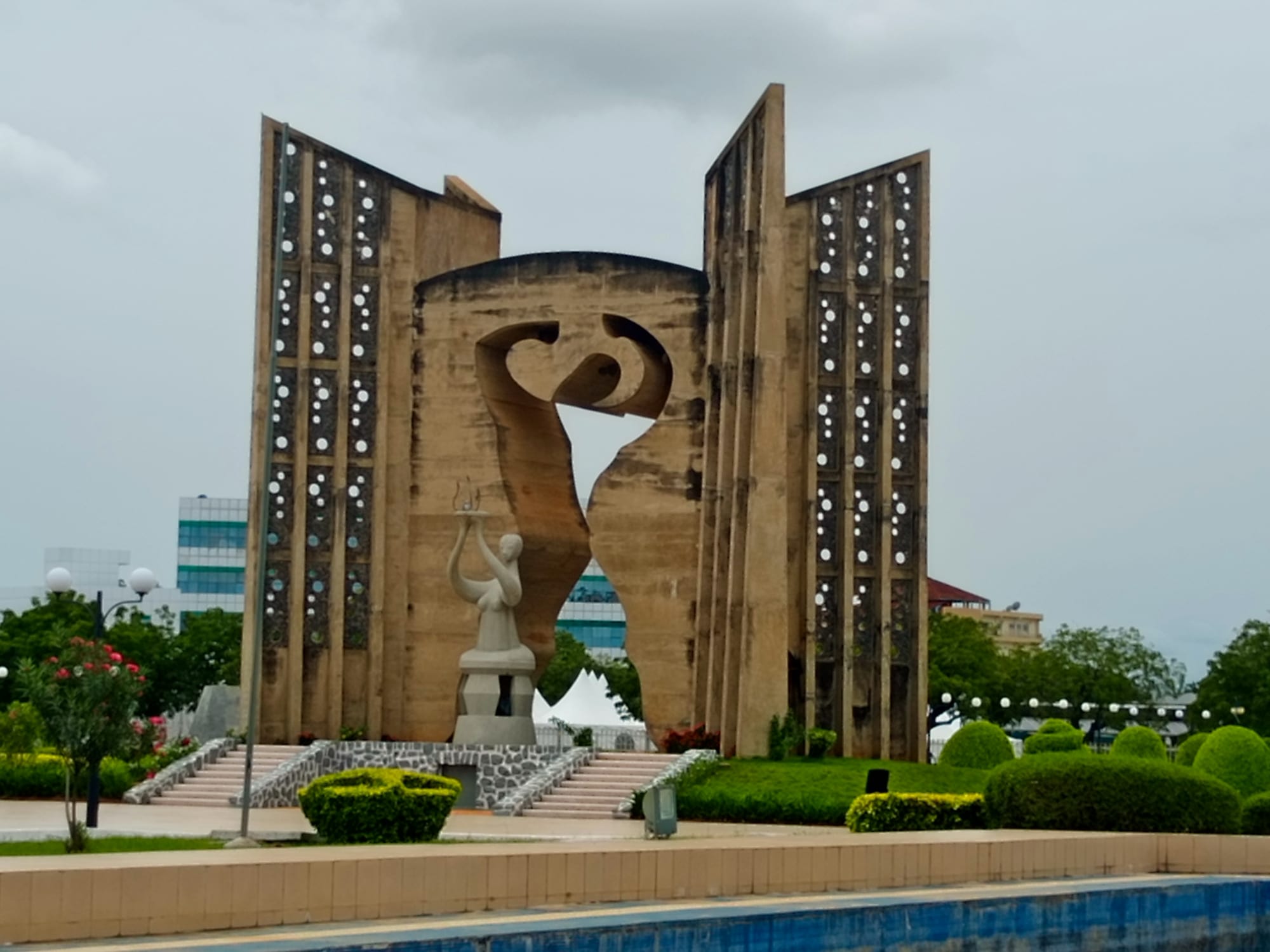 A tall concrete monument with the outline of a human figure cut through its centre, a pale statue holding a bowl aloft in front of it, set in clipped gardens