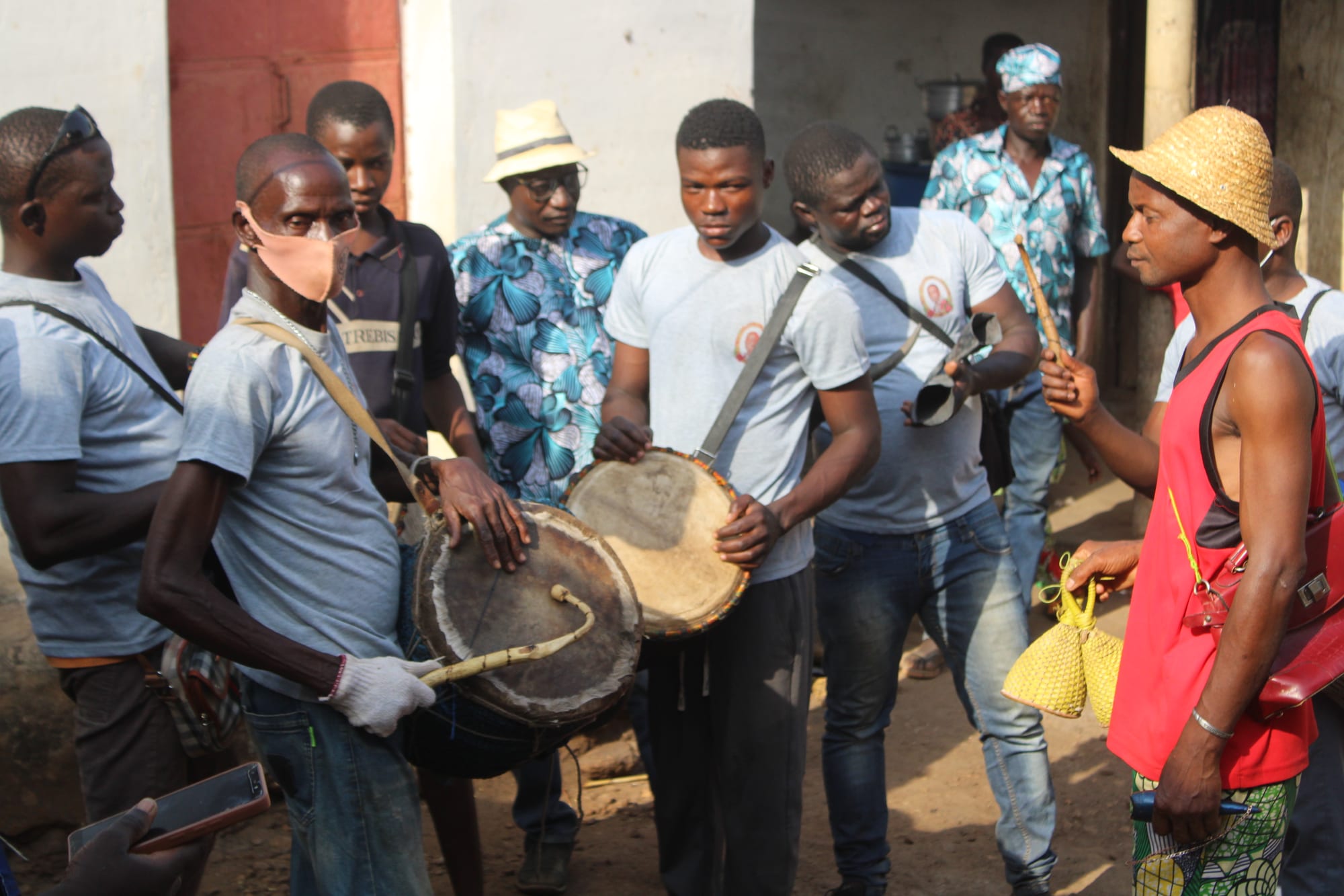 A group of men gathered in a dusty yard, two of them playing large hide-covered drums slung from shoulder straps while others hold rattles and a small bell