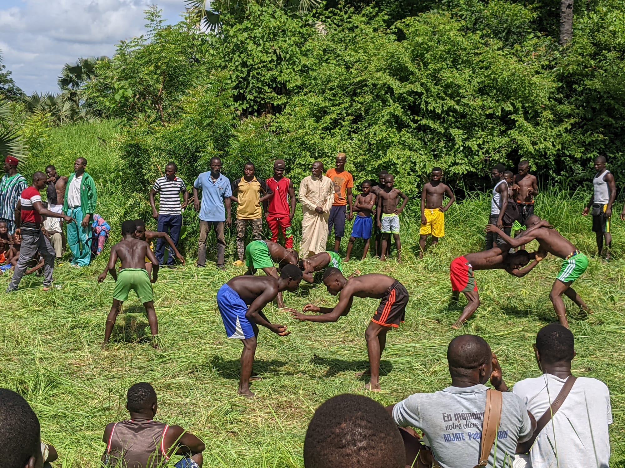 Pairs of bare-chested young men in shorts wrestling on a patch of trampled grass, watched by a ring of spectators in front of dense green bushes and palm trees