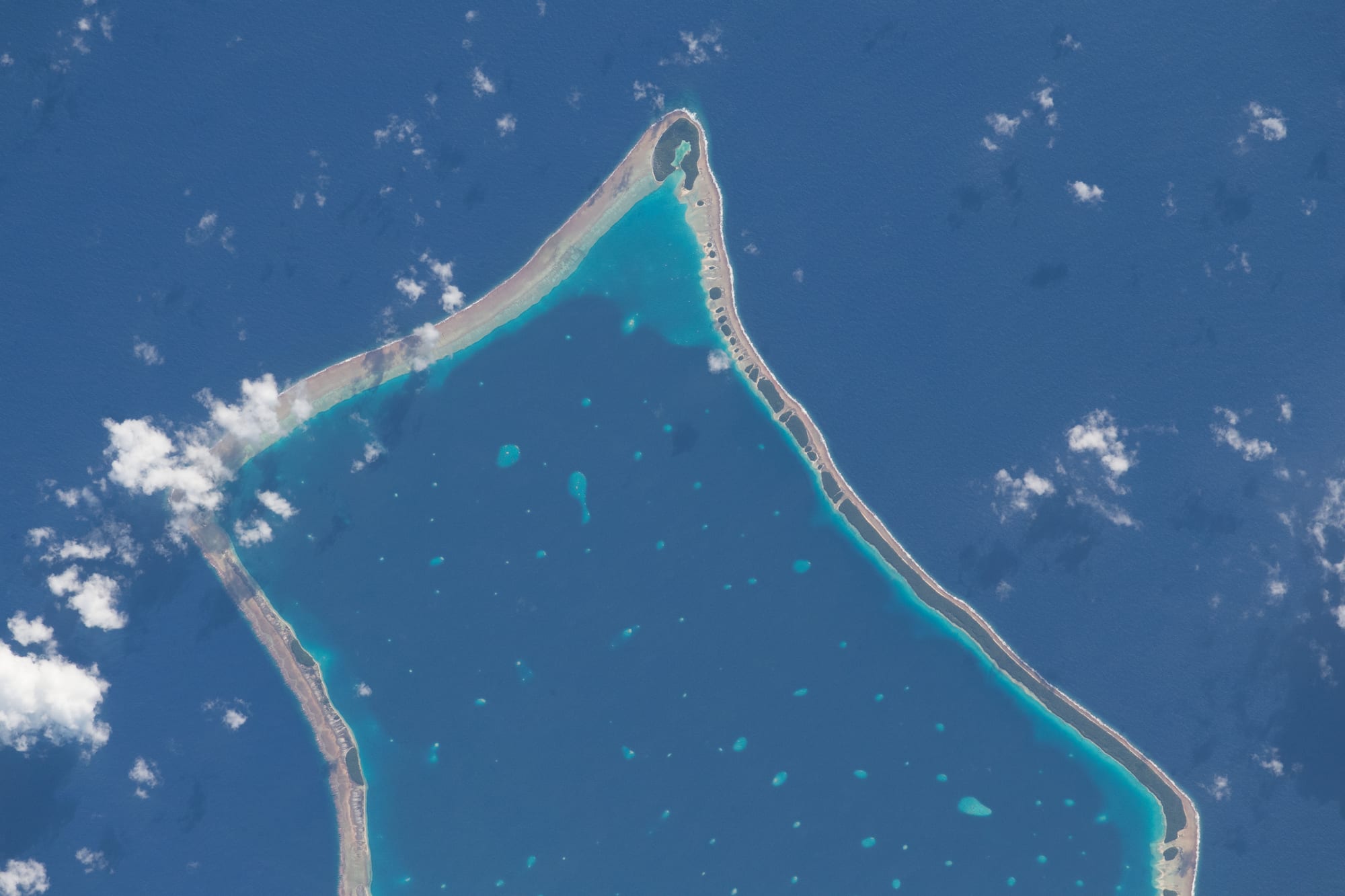 An aerial view of a coral atoll — a thin broken ring of green islets around a pale turquoise lagoon in the open ocean