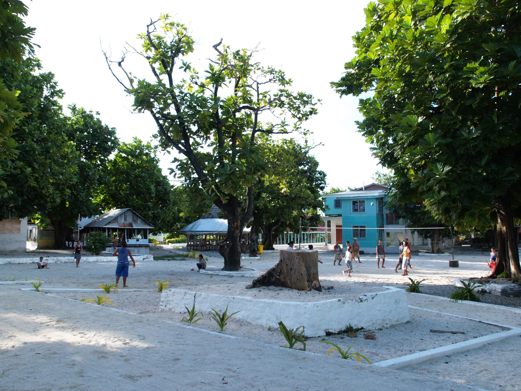 People playing volleyball on the open sandy village green of Fakaofo, surrounded by trees and simple colourful houses