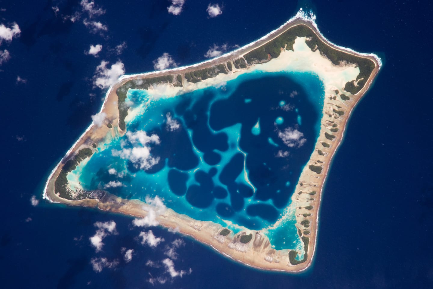 An aerial view of a narrow coral atoll ringed with palm trees surrounded by ocean