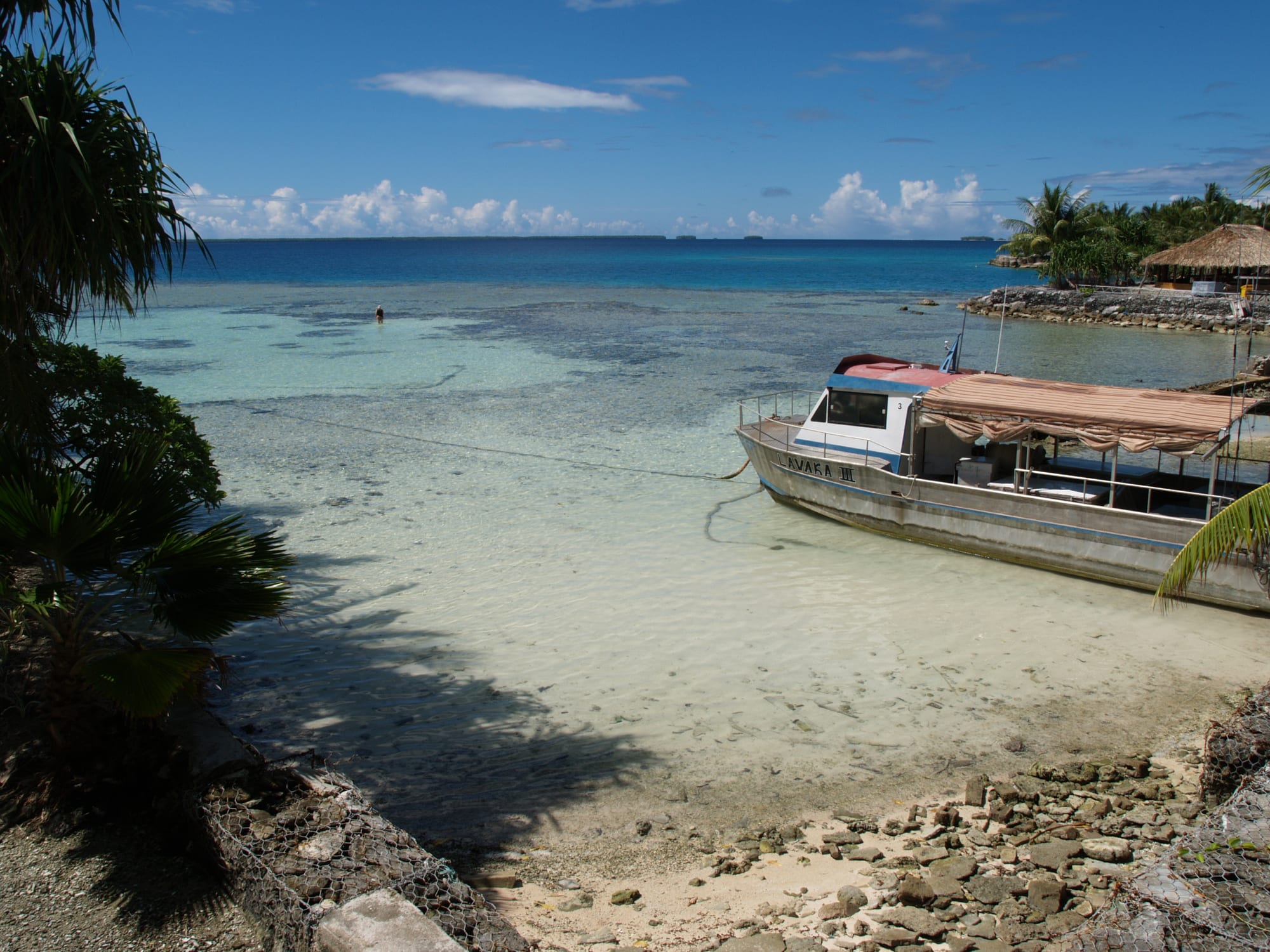 A shallow turquoise lagoon over pale coral sand, with a moored passenger boat on the right, a thatched shelter on a stone jetty and a line of low islets on the horizon
