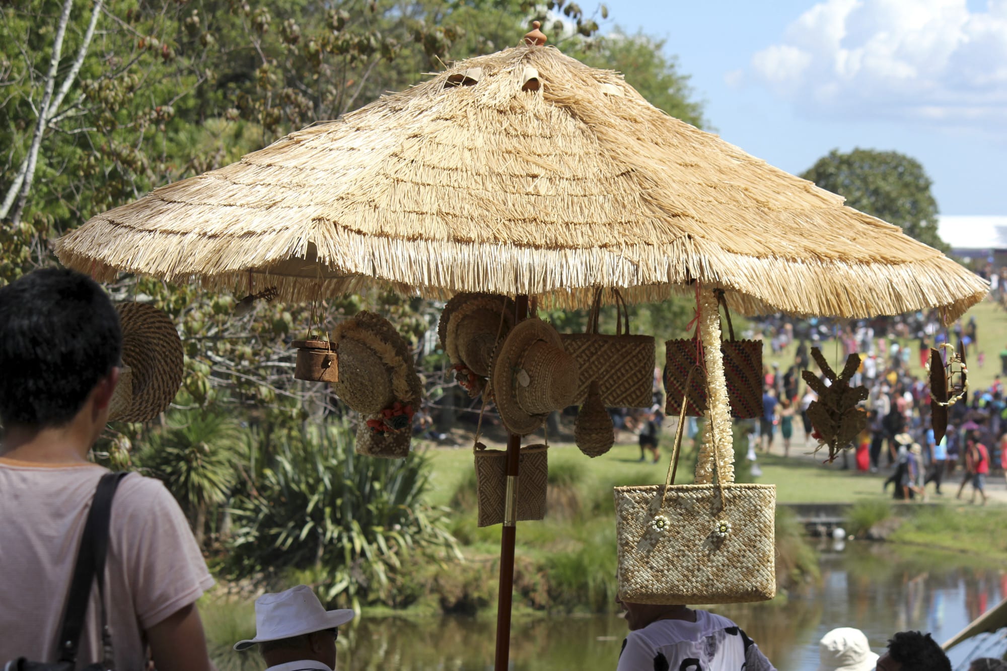 Hand-woven bags, hats and baskets hanging from a large thatched umbrella at an outdoor festival, with a crowd on a grassy slope beyond