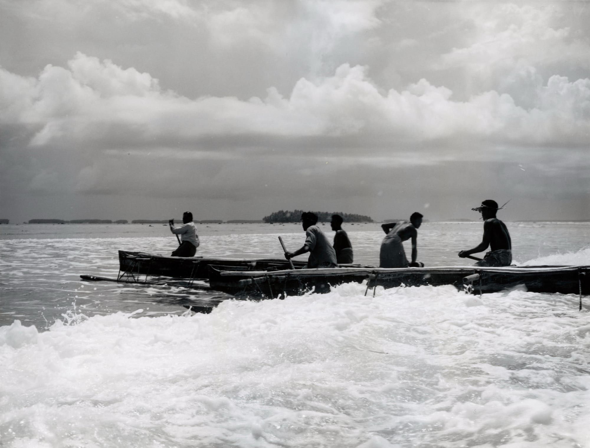 A black and white photograph of men paddling long canoes through white surf over a reef, with a low wooded islet on the horizon and heavy clouds above
