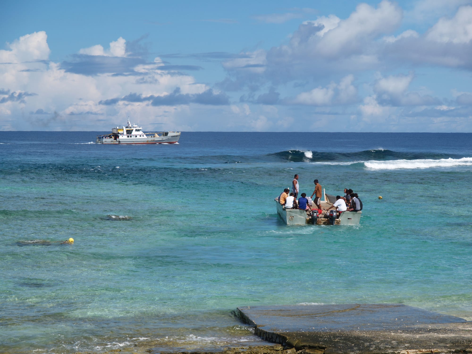An open metal barge carrying a group of people heads out across shallow turquoise water from a concrete ramp towards a white ship waiting beyond the breaking surf on the reef