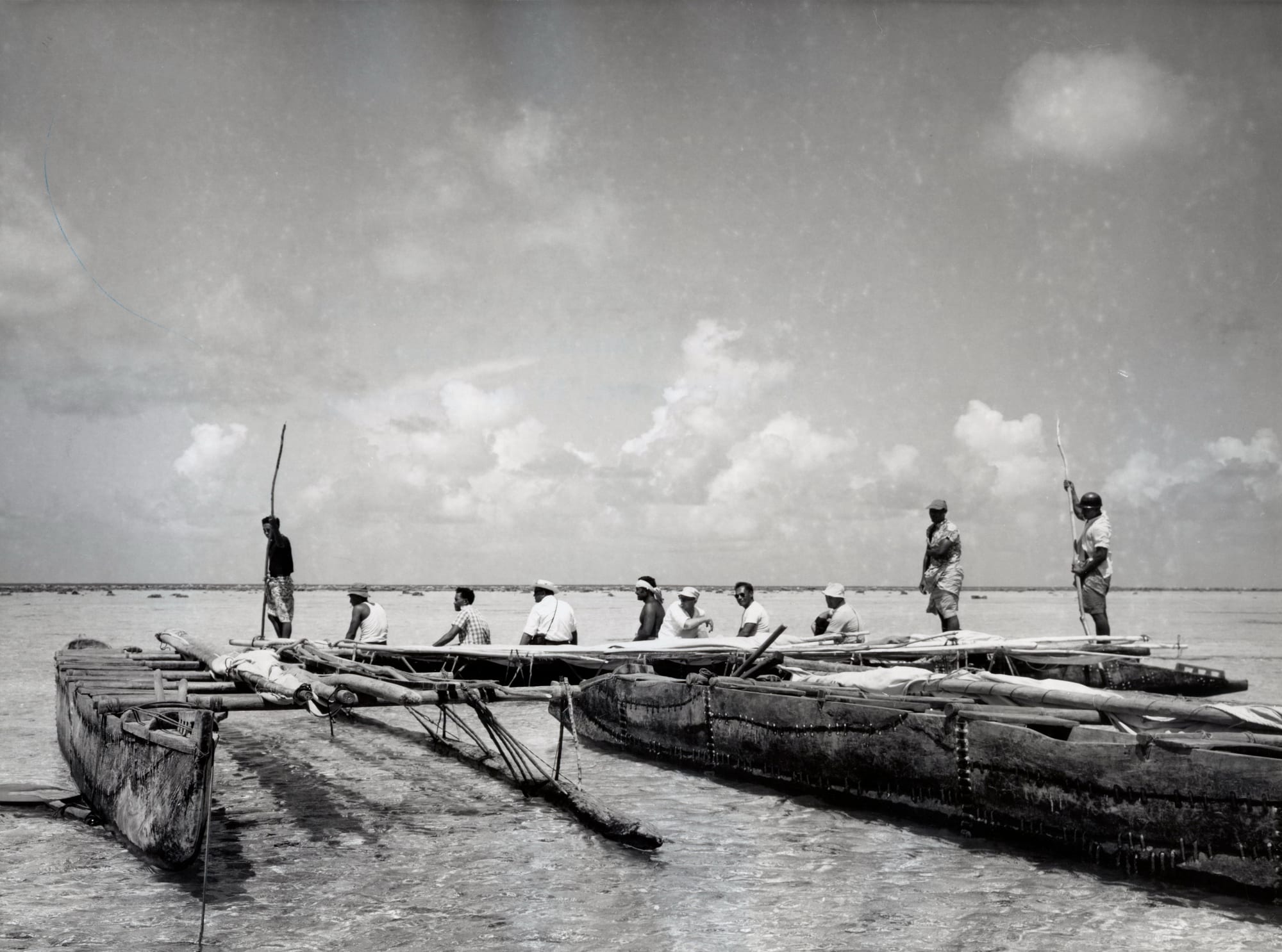 A black-and-white photograph of wooden outrigger canoes in shallow water, with a row of men in hats sitting in one and two men standing with poles