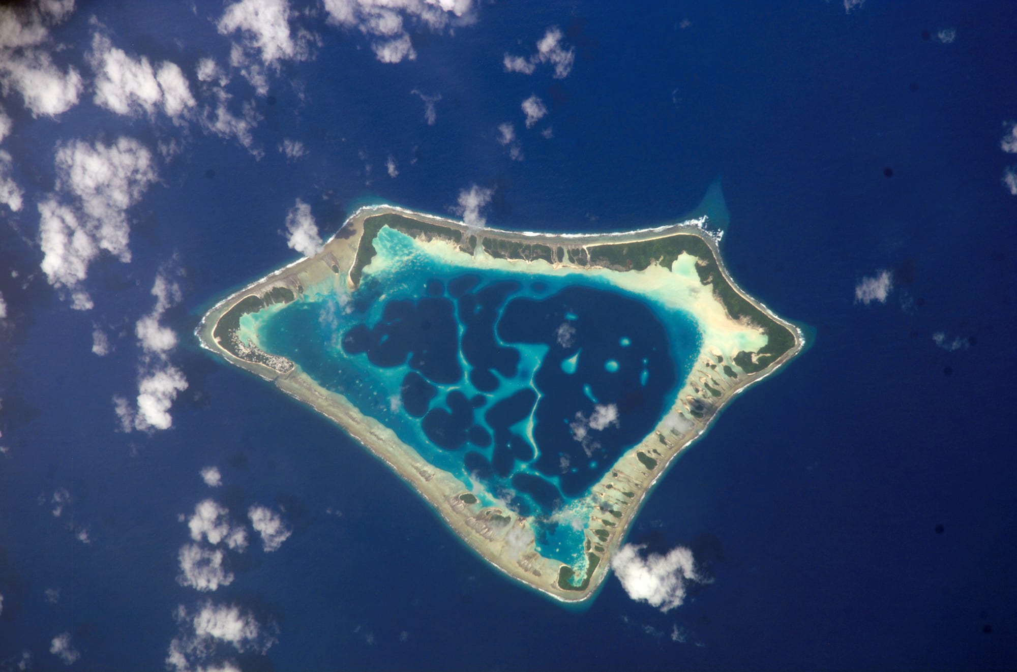 An astronaut photograph of a narrow ring-shaped coral atoll enclosing a turquoise lagoon in dark ocean