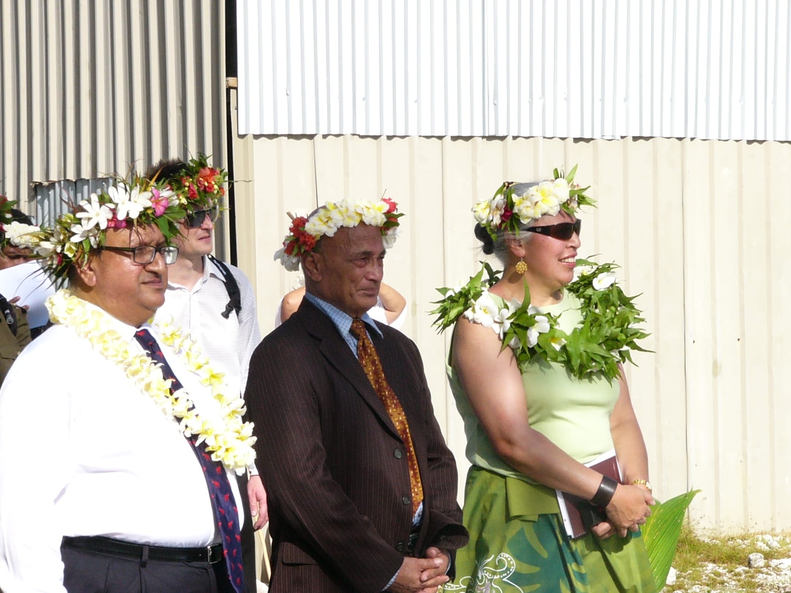 Two men and a woman wearing flower garlands and head wreaths standing in front of a corrugated-iron building