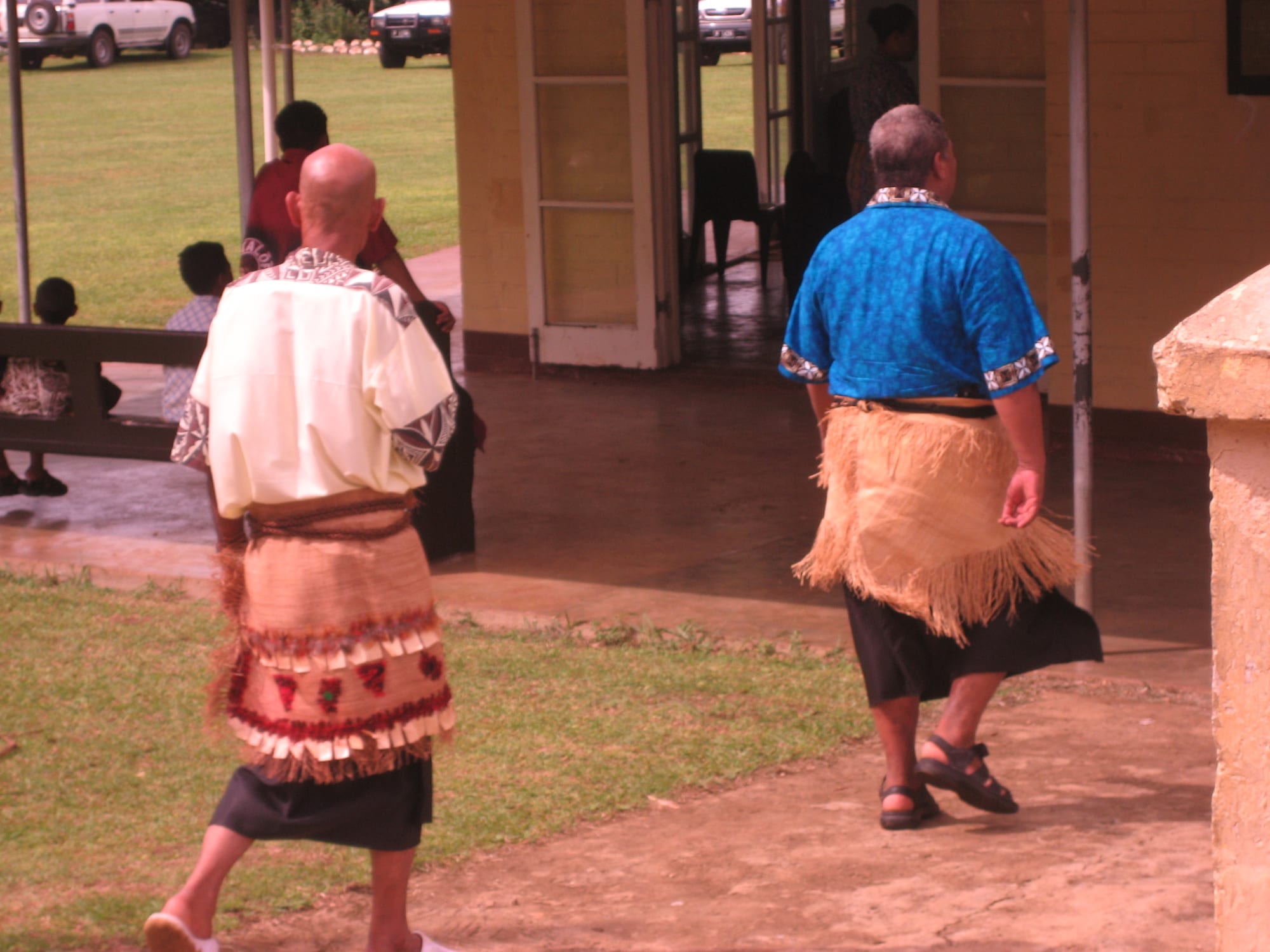 Two men seen from behind walk across a wet path towards a building, both wearing woven fibre mats tied around the waist over dark skirts, one in a cream shirt and one in a bright blue shirt