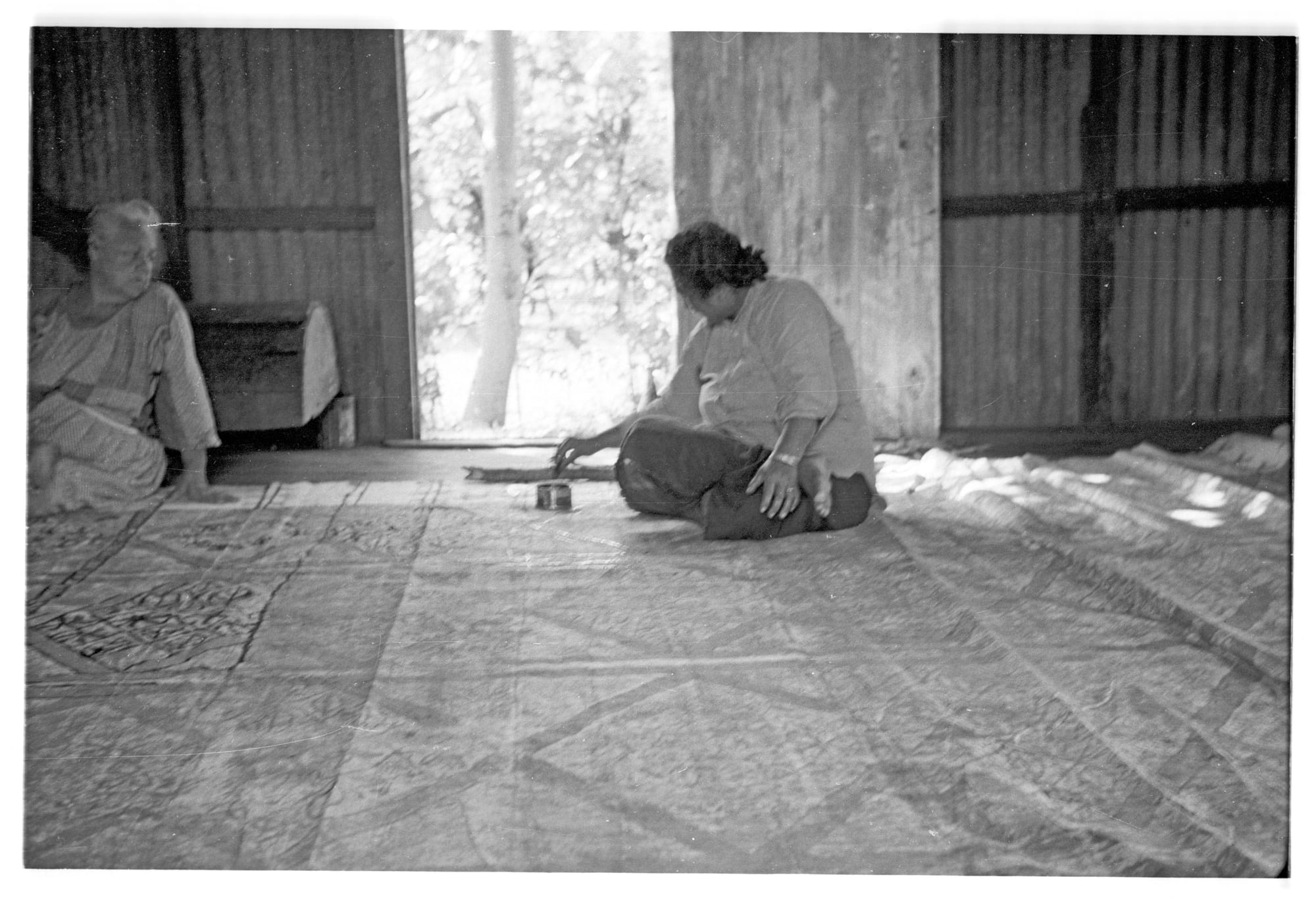 A black and white photograph of a woman sitting on a large sheet of patterned bark cloth spread across a wooden floor, painting it by hand, while an older woman sits nearby at the left