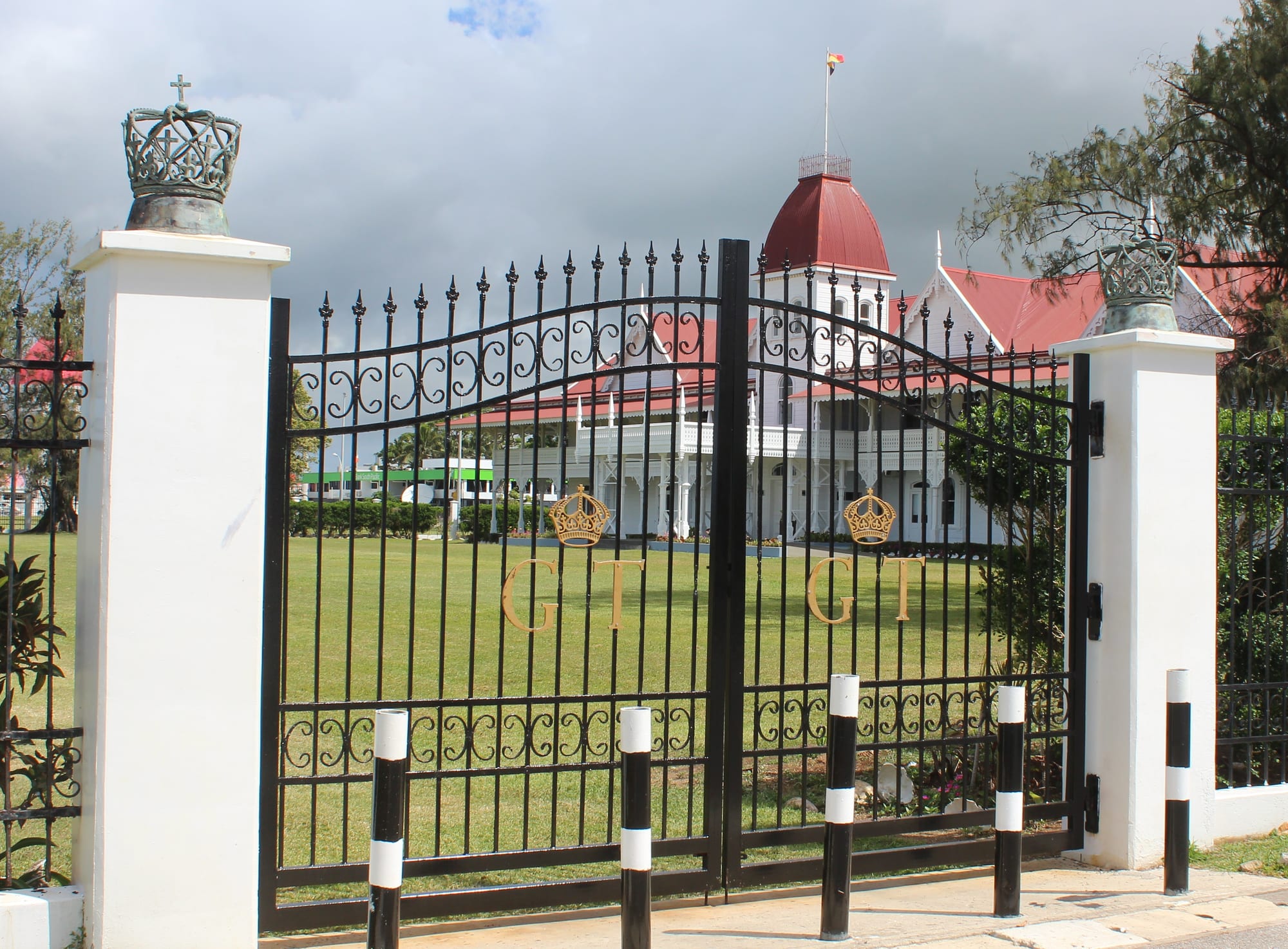Black iron gates decorated with gold crowns and the letters G and T, set between white pillars topped with crowns, in front of a lawn and a white wooden palace with a red roof and a red-domed tower