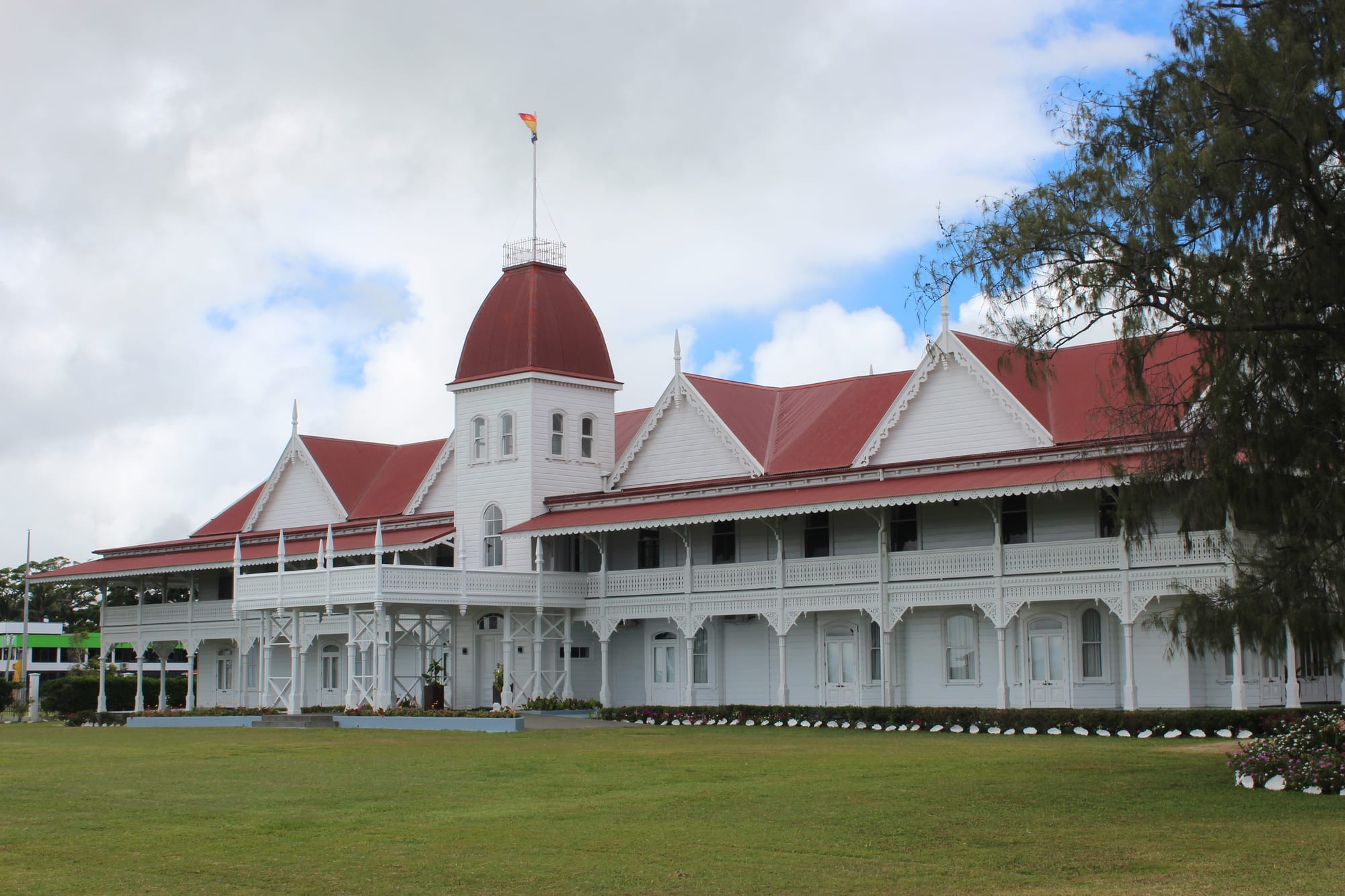 A white Victorian timber palace with red roofs and verandahs behind a lawn and Norfolk pines in Nukuʻalofa