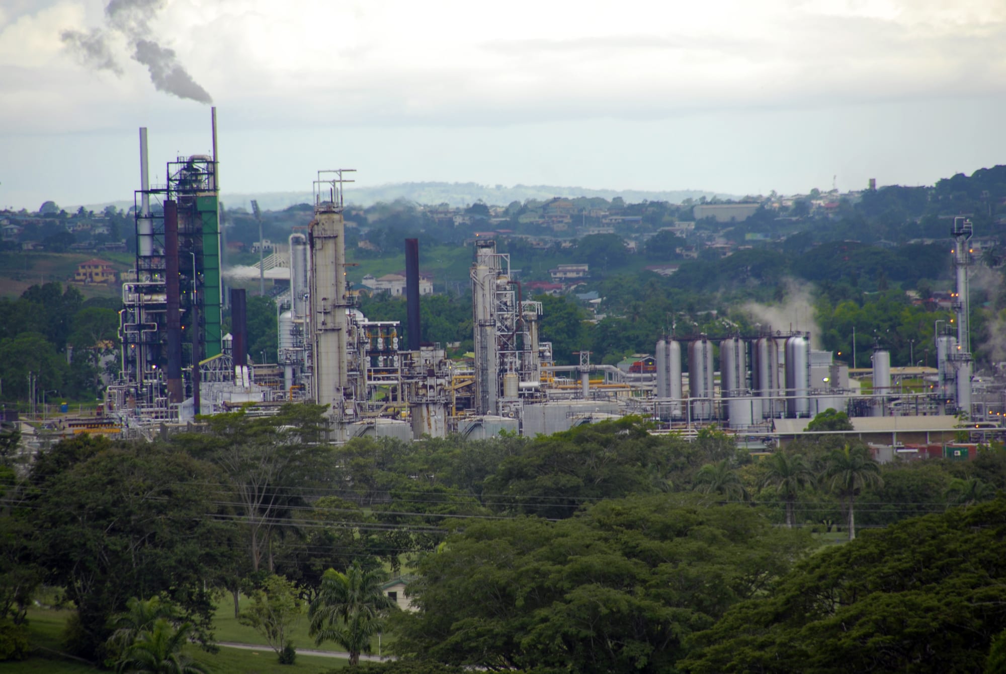 Steel towers, tanks and chimneys of an oil refinery rising above green trees, with a plume of smoke and low hills behind
