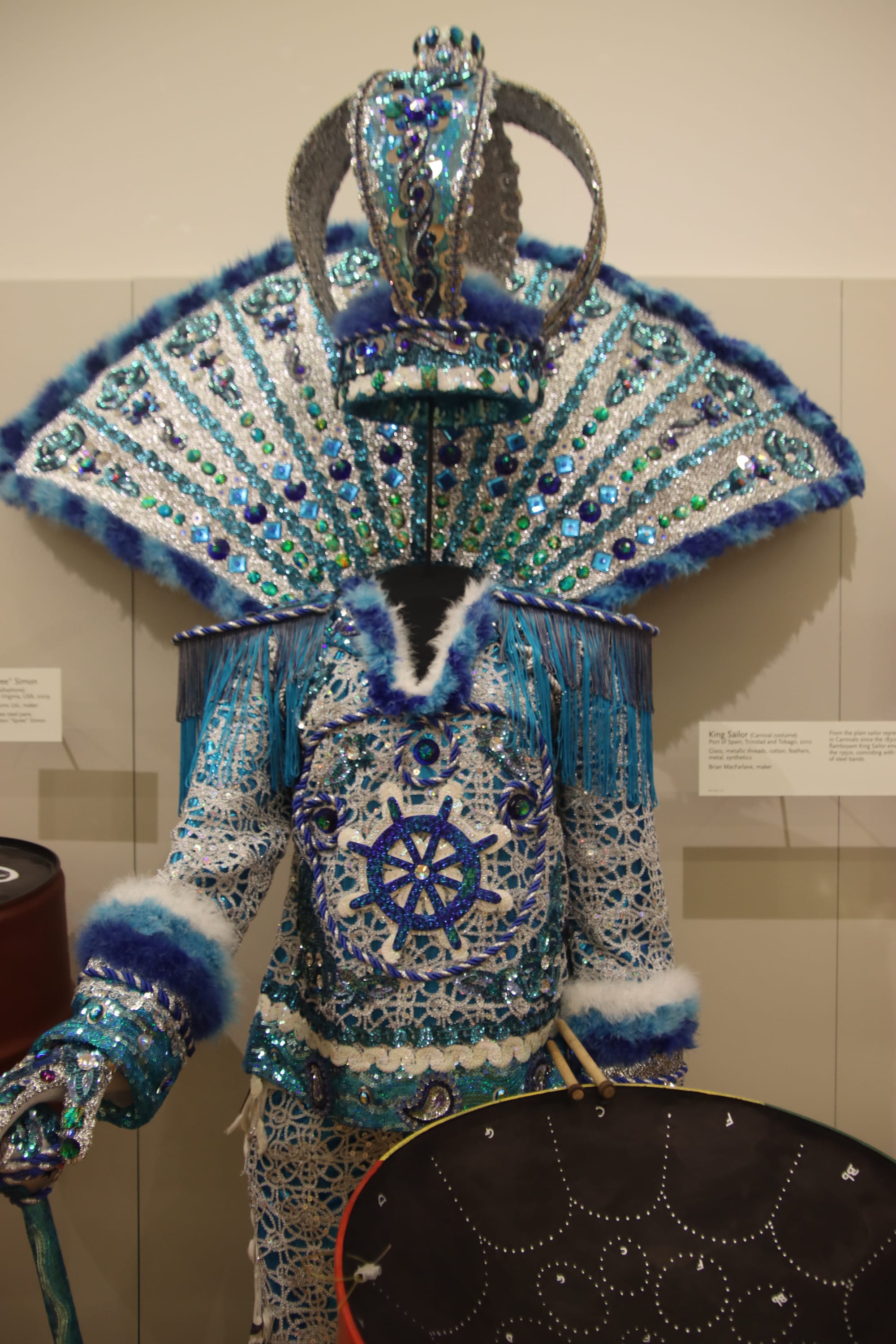 An elaborate carnival costume of blue and silver sequins, beads and feathers, with a tall crown and a fan-shaped back piece, displayed on a stand behind a steelpan