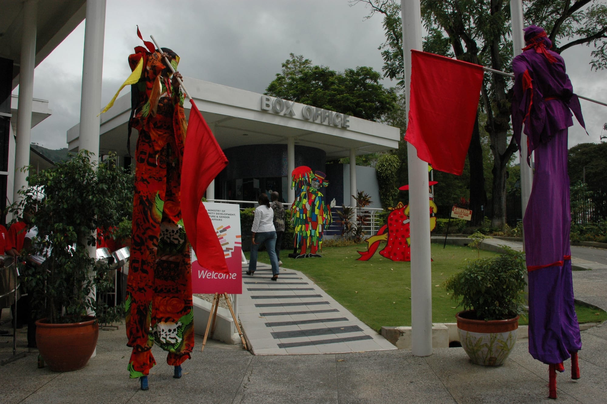 Two stilt walkers in long flowing costumes, one in red patterned cloth holding a red flag, the other in purple with a red sash, stand either side of a path leading to a building marked Box Office, with more costumed figures on the lawn behind
