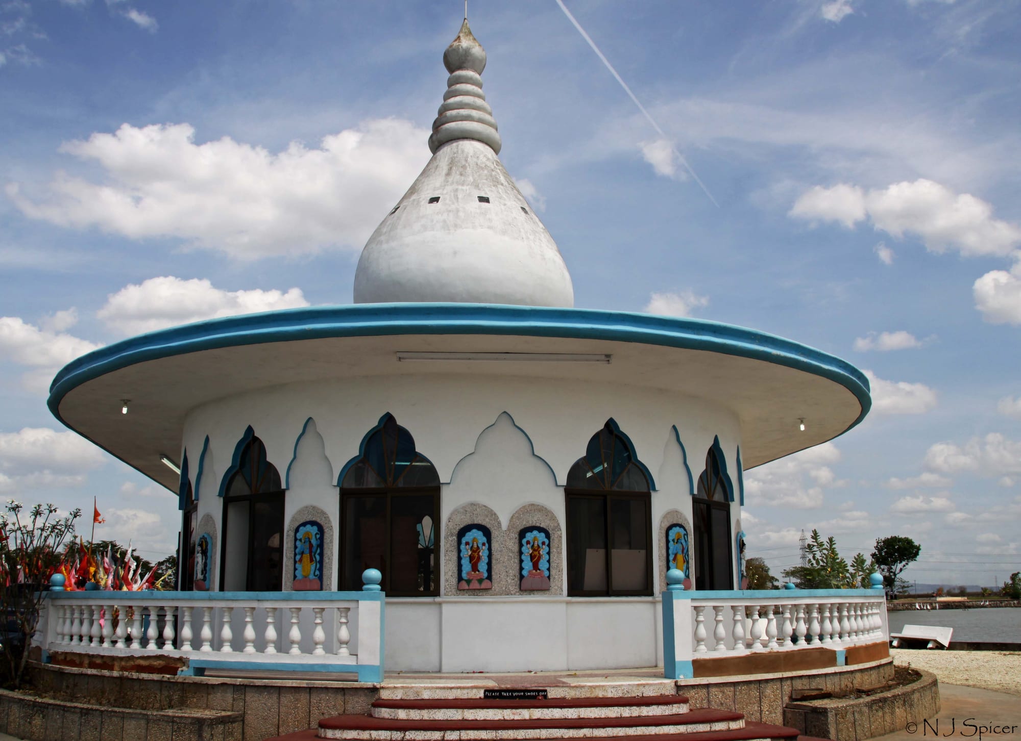 A round white Hindu temple with a tall onion-shaped dome and a wide blue-edged roof, arched windows set with painted images of deities, a white balustrade and steps in front, and coloured flags at the left