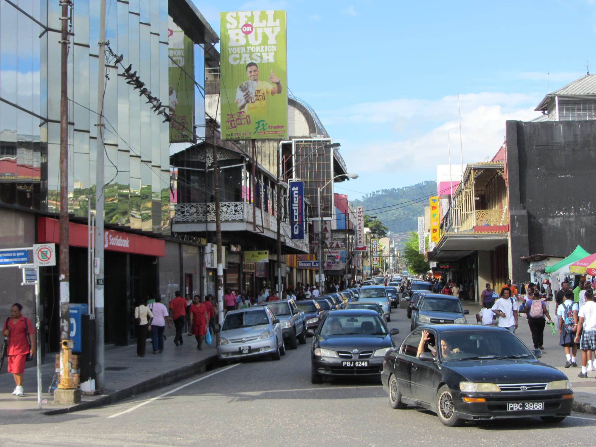 A busy downtown street in Port of Spain with cars queued along the road and pedestrians, including schoolchildren, walking past shops