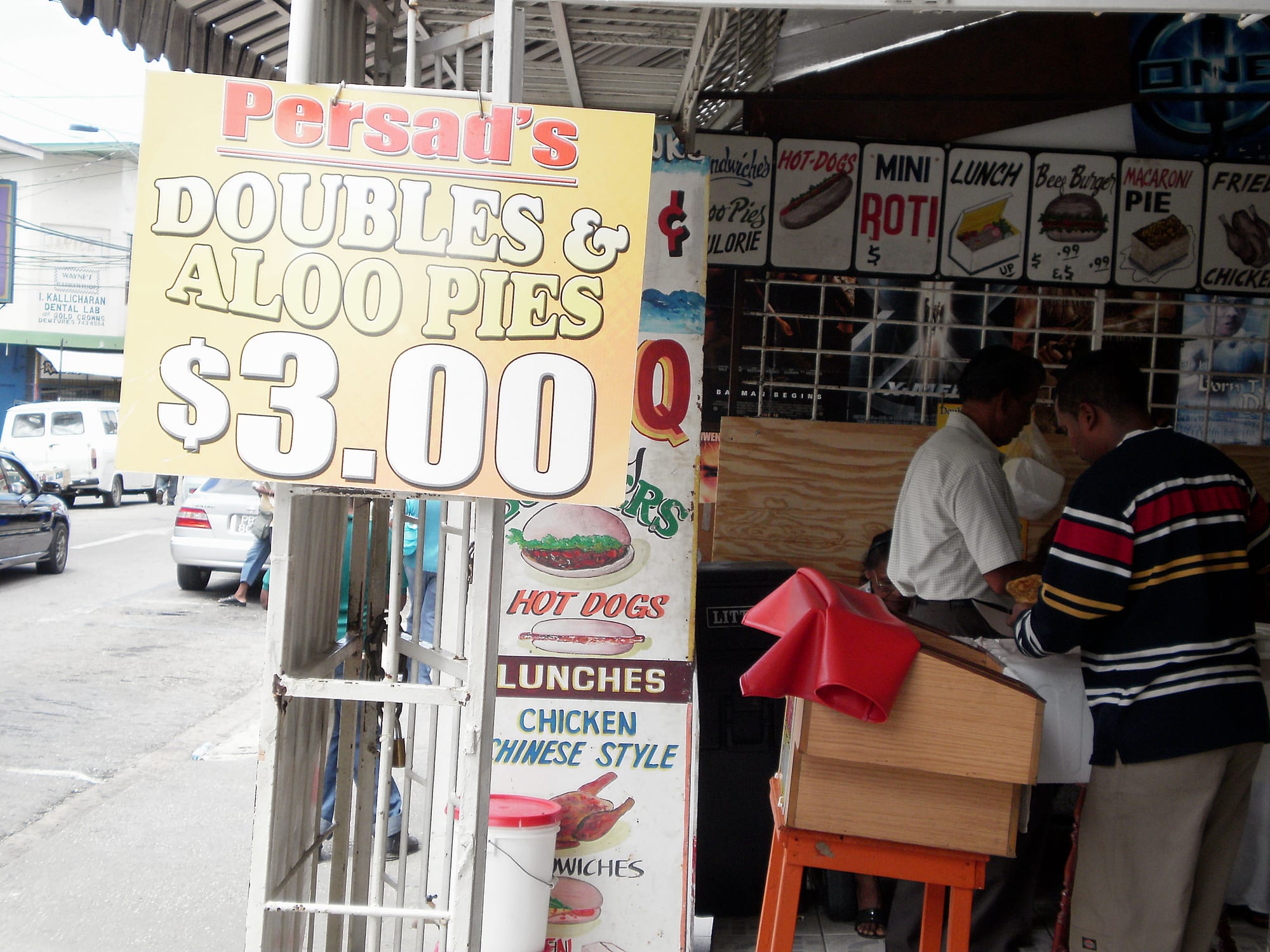 A roadside doubles and aloo pie stand with a large price sign, customers being served at the counter