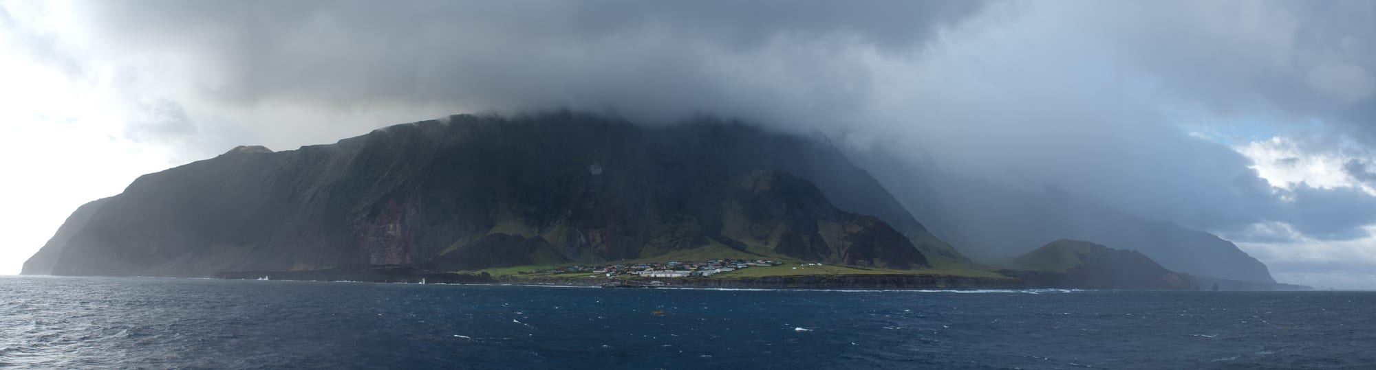 A small settlement beneath a towering volcanic peak with a storm approaching