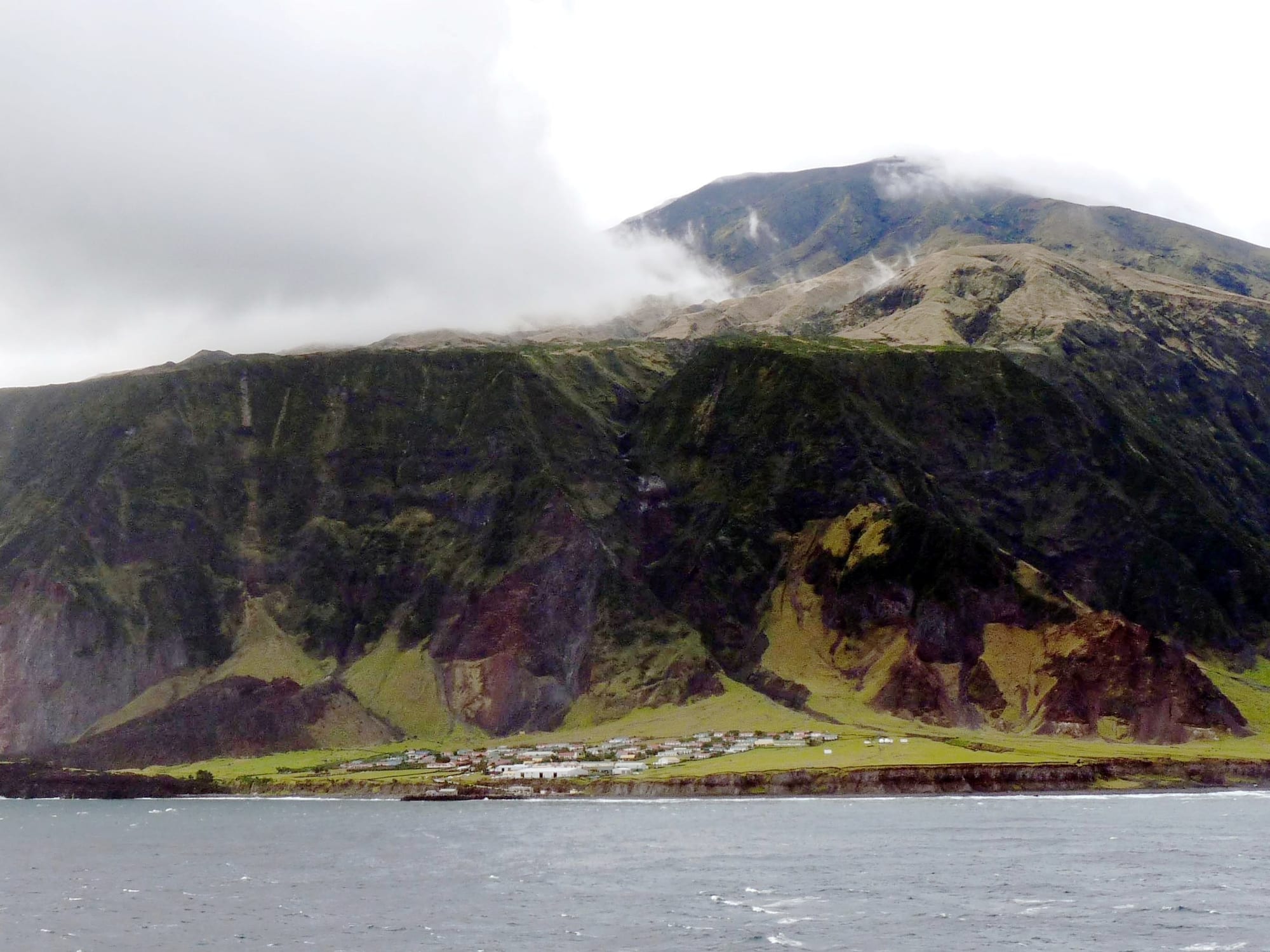 A small cluster of white houses on a narrow green coastal plain at the foot of steep, dark volcanic cliffs, with cloud drifting over the upper slopes and grey sea in the foreground