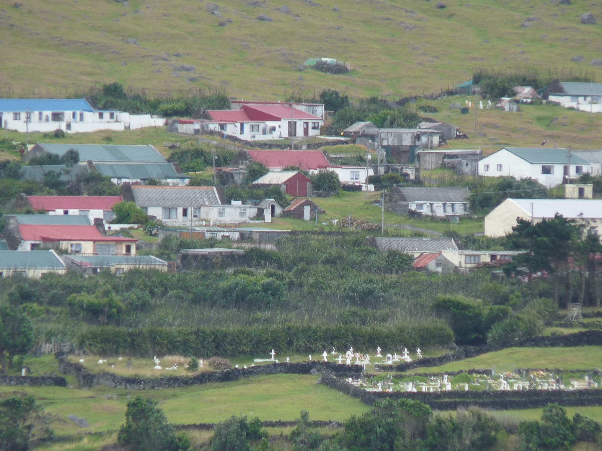 Houses with red, blue and grey roofs spread across a green hillside, with stone walls and rows of white crosses and headstones in a cemetery in the foreground