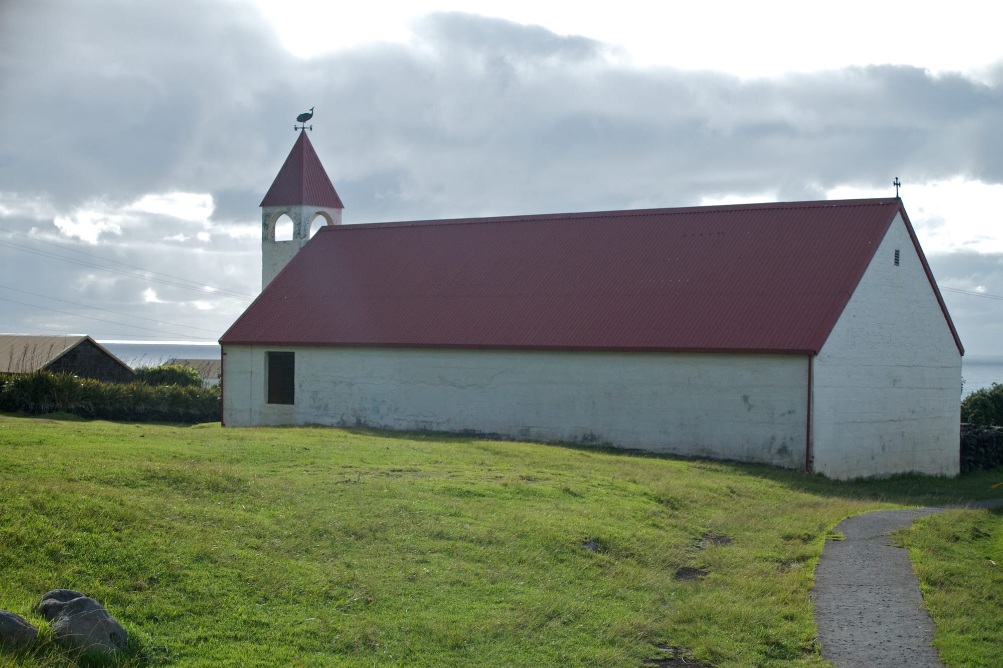 A long, low whitewashed church with a red corrugated roof and a small square bell tower topped by a weather vane, standing on a grassy slope with a paved path in front