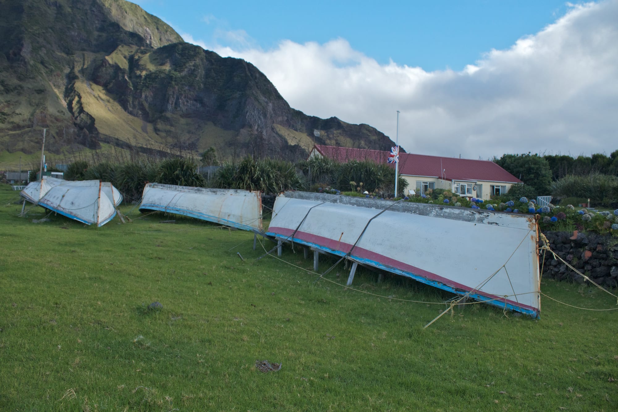 Three white canvas-covered longboats with coloured trim resting upside down on wooden supports on a lawn, with a red-roofed house, a flagpole and steep green cliffs behind