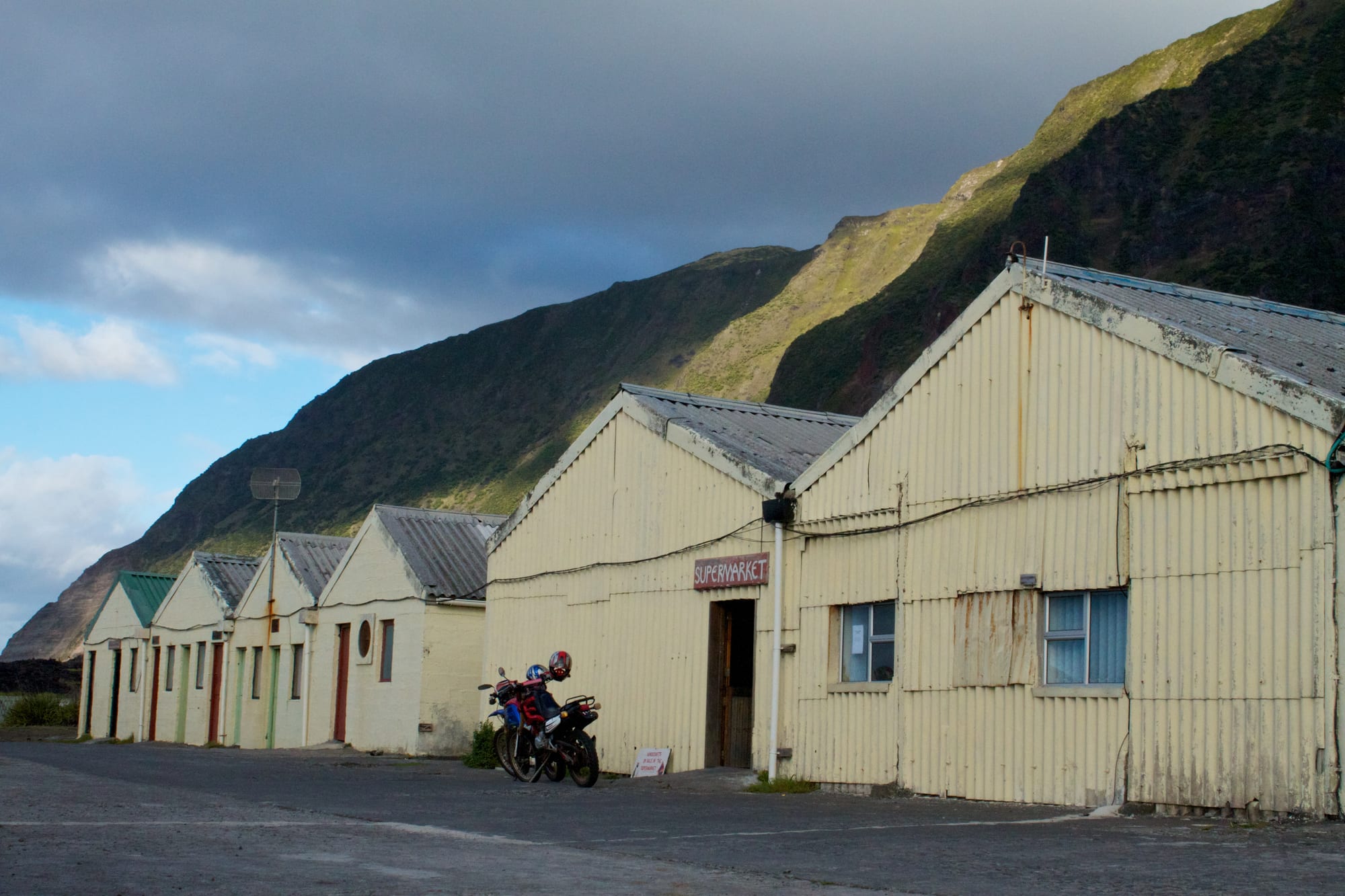 A corrugated-iron building with a hand-painted 'Supermarket' sign, motorbikes parked outside, beneath a steep green hillside
