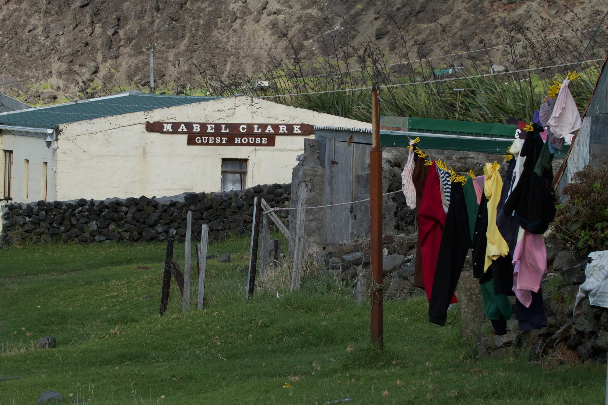 A row of laundry hanging on a line in a grassy yard in front of a whitewashed cottage with a sign reading Mabel Clark Guest House, below a steep volcanic slope