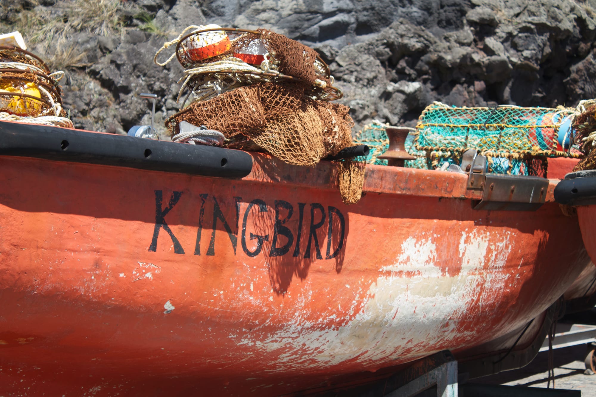 The bow of an orange fishing longboat named Kingbird, piled with rope and lobster traps, against a backdrop of black volcanic rock