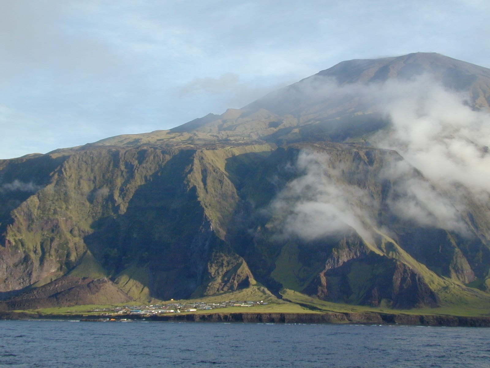 Rows of low houses and green fields of the Tristan da Cunha settlement below dark volcanic cliffs