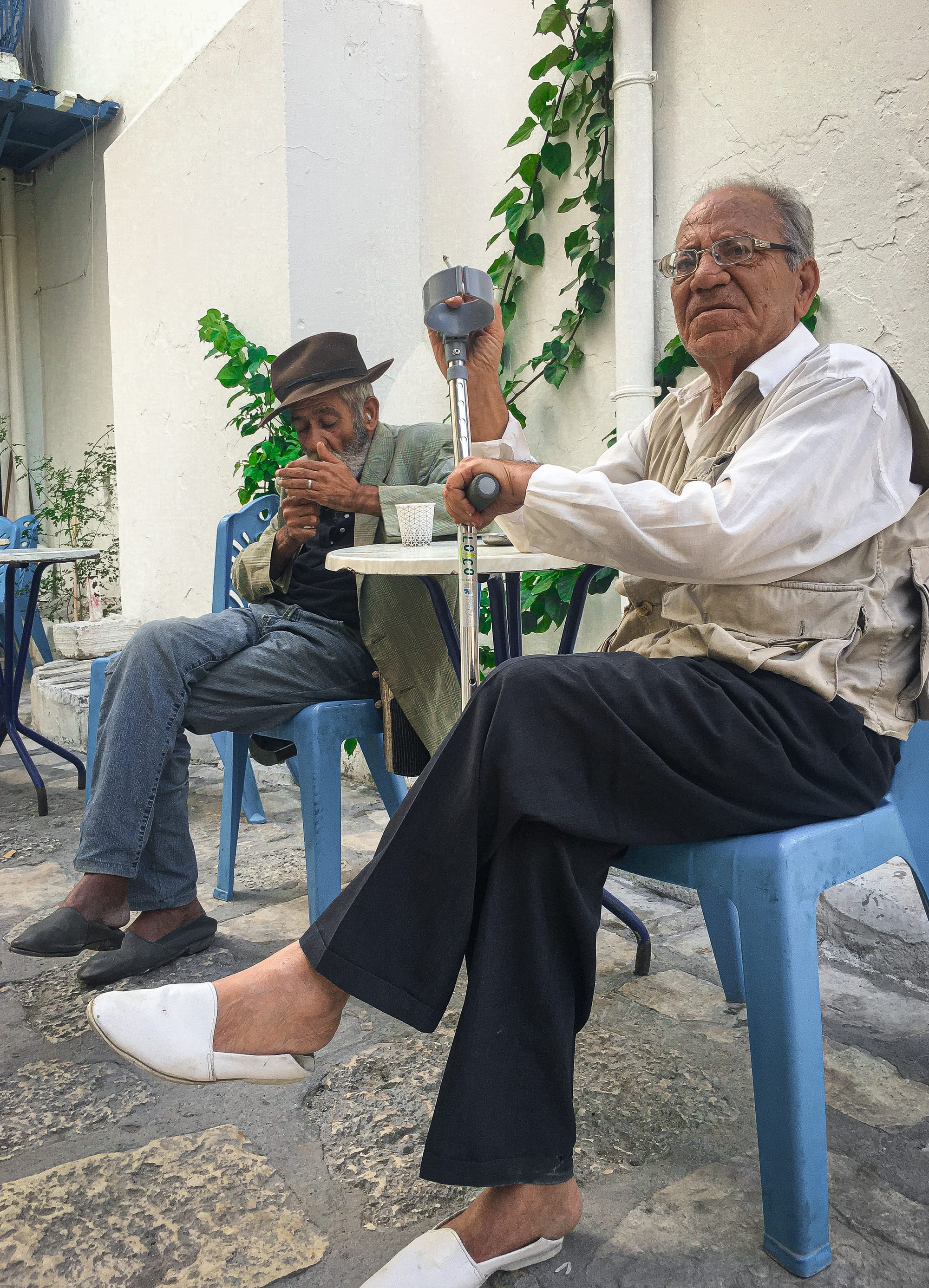 Two older men sitting at a small café table in a medina courtyard, one lighting a cigarette, the other resting a hand on his cane