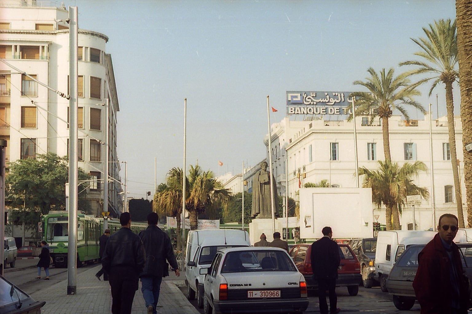 A tree-lined boulevard in Tunis with a tram, parked cars and pedestrians walking past white colonial-era buildings