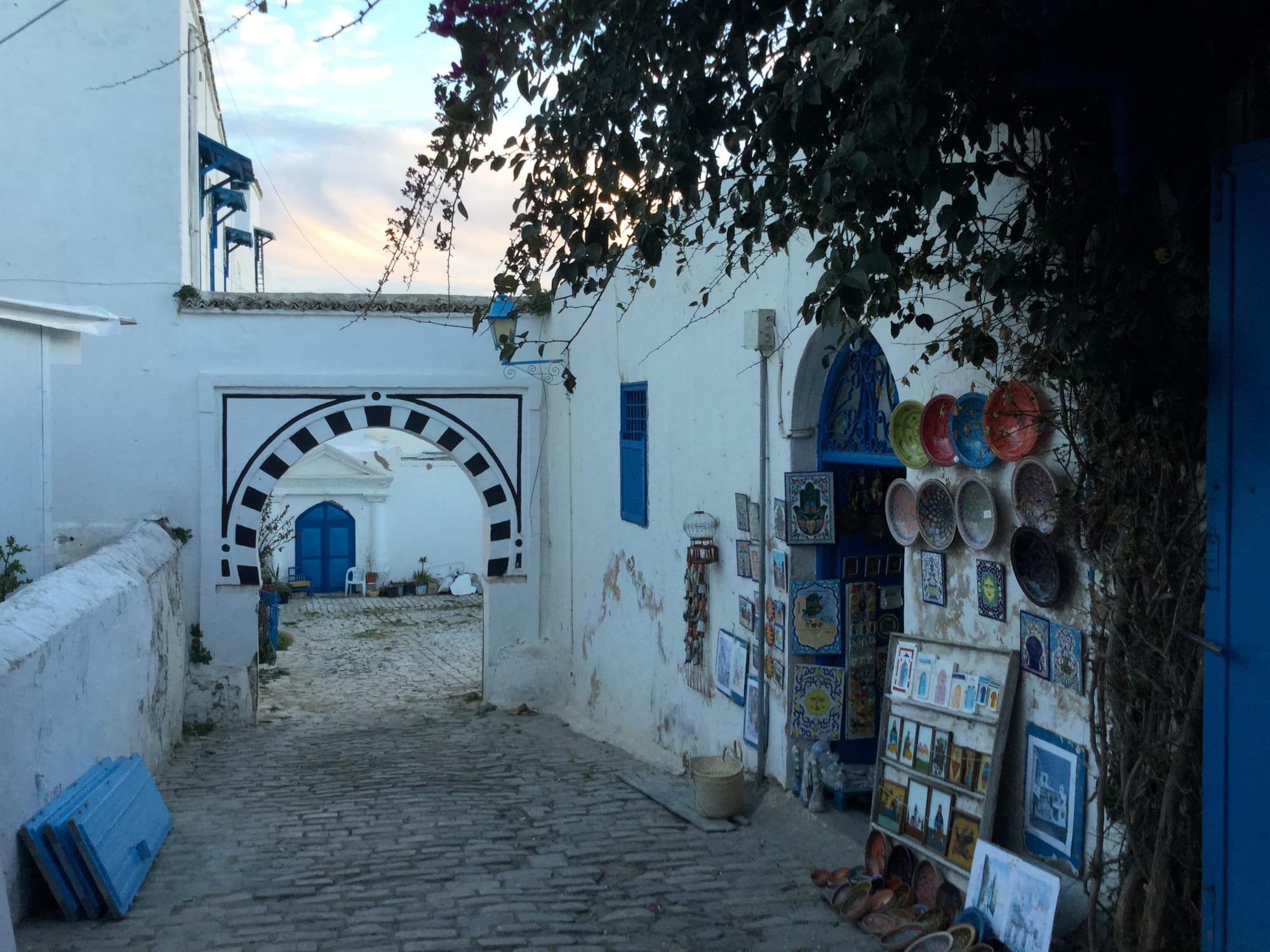 A cobbled alley in Sidi Bou Said between whitewashed walls with blue shutters, a black-and-white arched gateway ahead