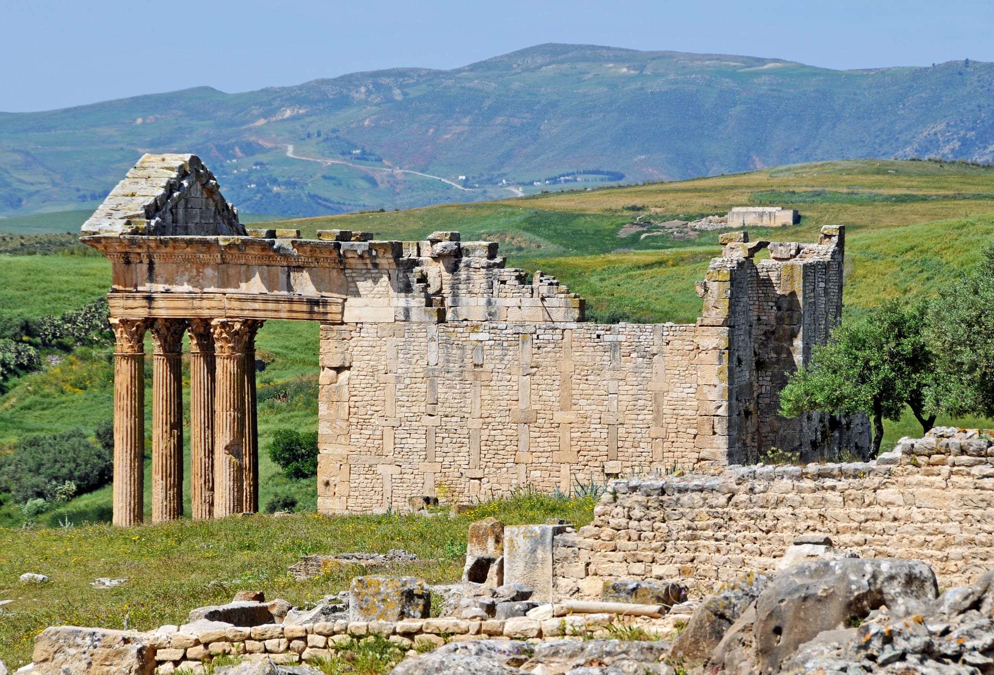 The columned porch and stone walls of a ruined Roman temple standing among green fields, with rolling farmland and hills behind