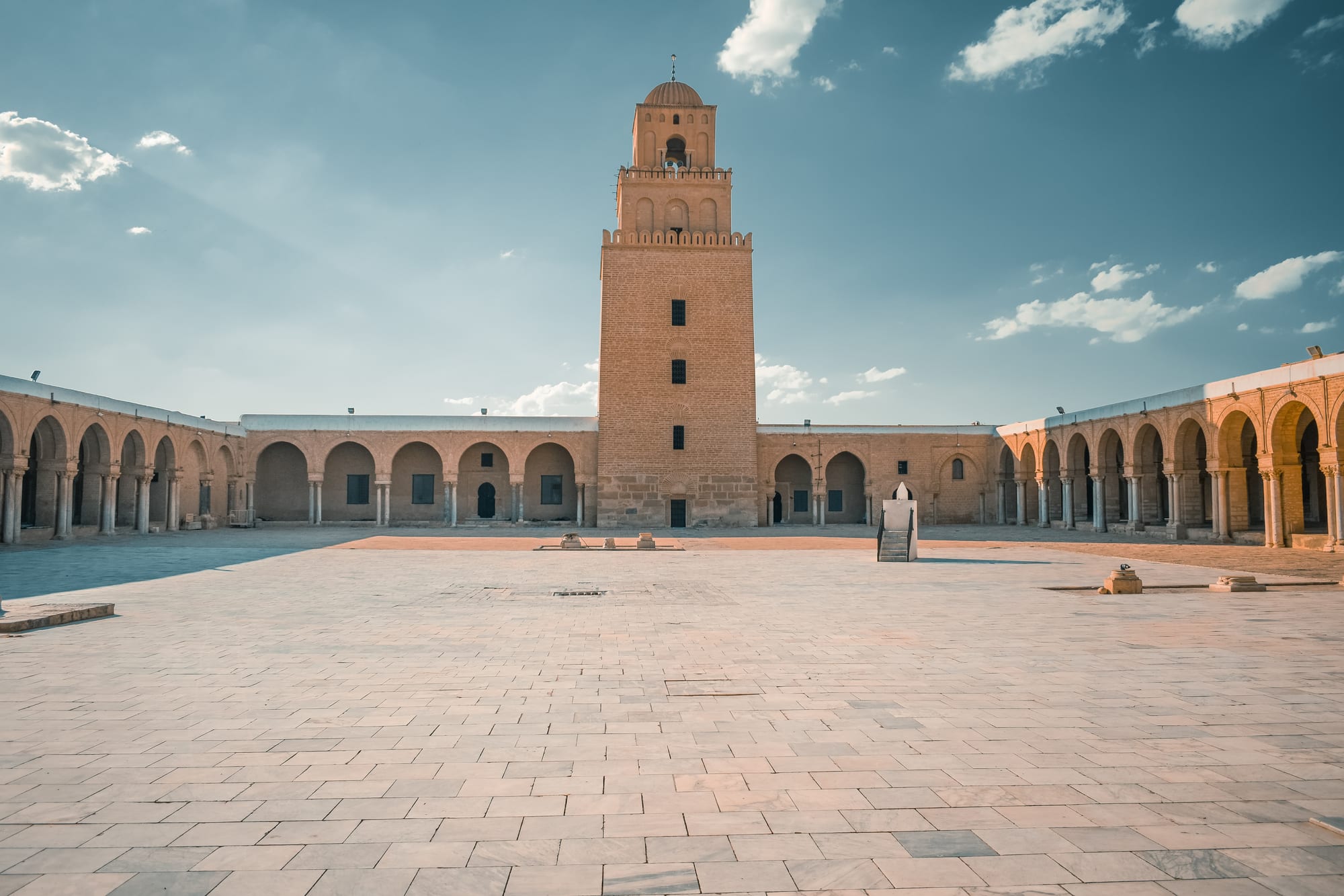 A vast paved courtyard surrounded by arcades of horseshoe arches, with a tall, square, three-tiered stone minaret rising at the far end under a blue sky