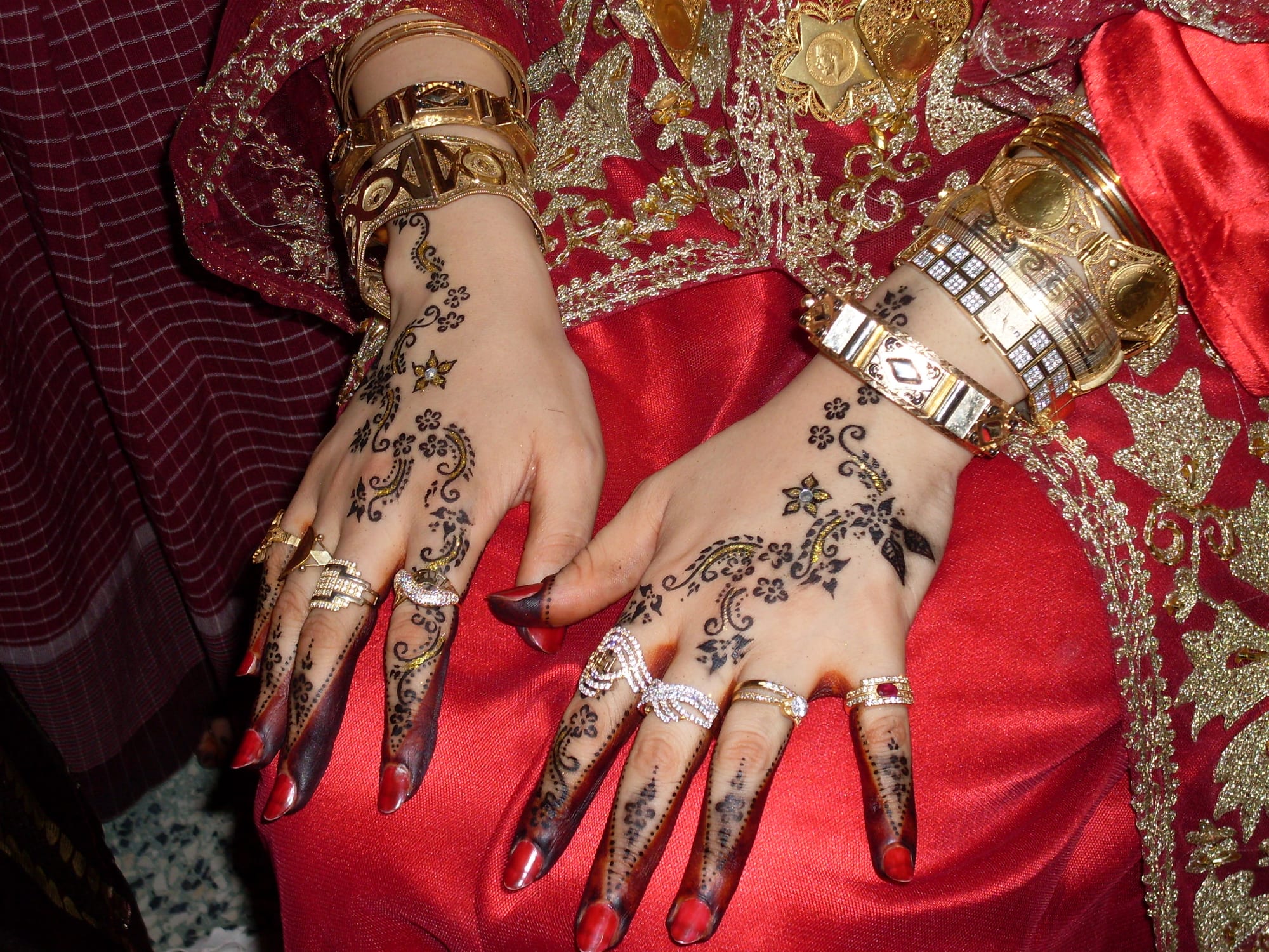 A woman's hands decorated with dark henna patterns of flowers and swirls, her fingertips dyed, wearing gold bangles and rings and resting on red cloth embroidered with gold
