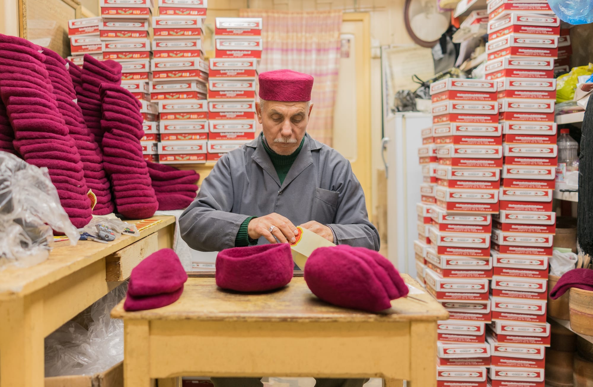 An older man in a grey work coat and a red felt cap sits at a wooden table finishing a red chechia, with stacks of red caps on one side and towers of red and white boxes behind him