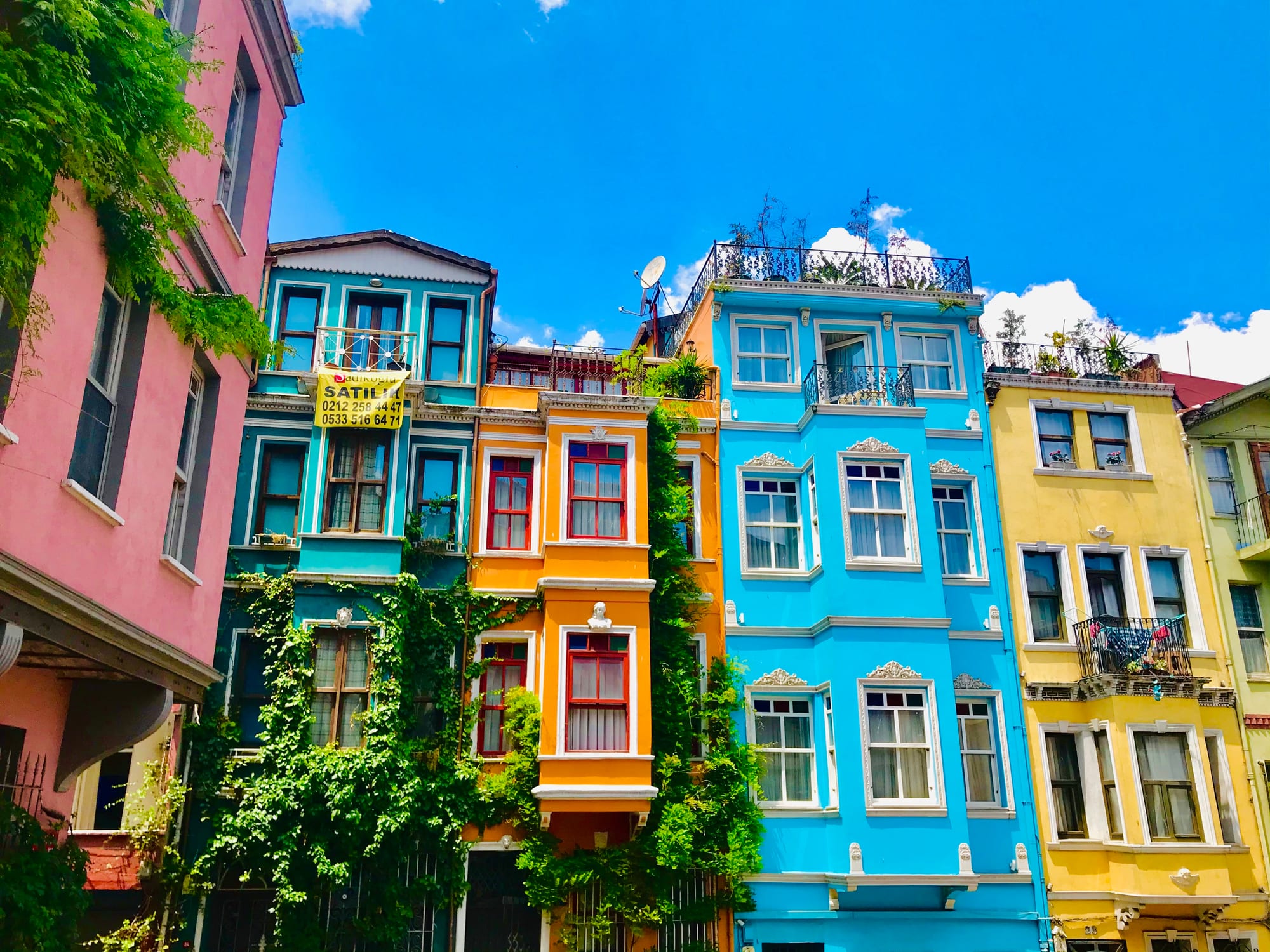 Rows of narrow apartment buildings painted pink, teal, orange, blue and yellow, with balconies and climbing plants, against a blue sky