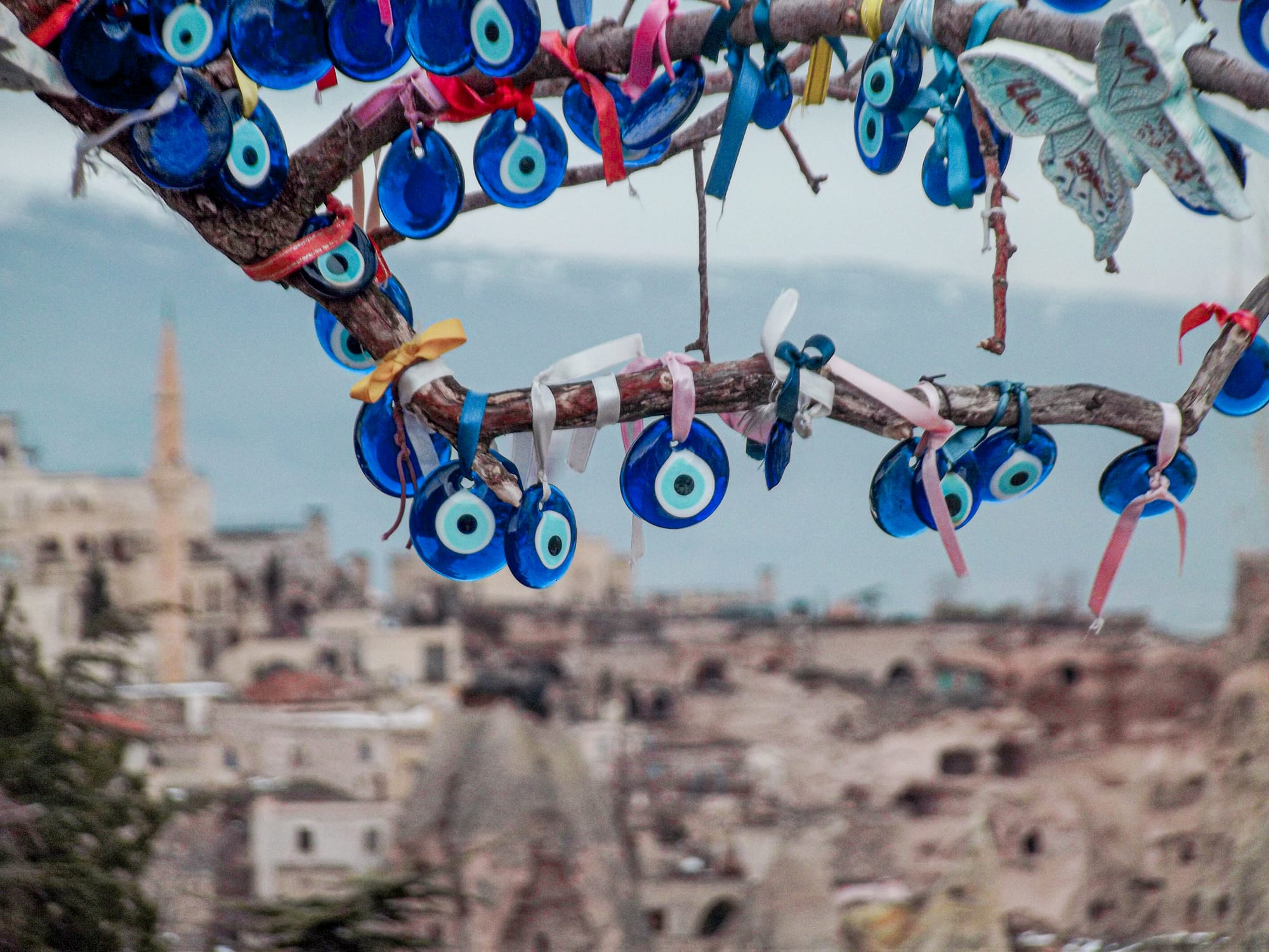 Dozens of round blue and white glass eye charms tied with coloured ribbons hang from the branches of a tree, with a town and a minaret blurred in the background