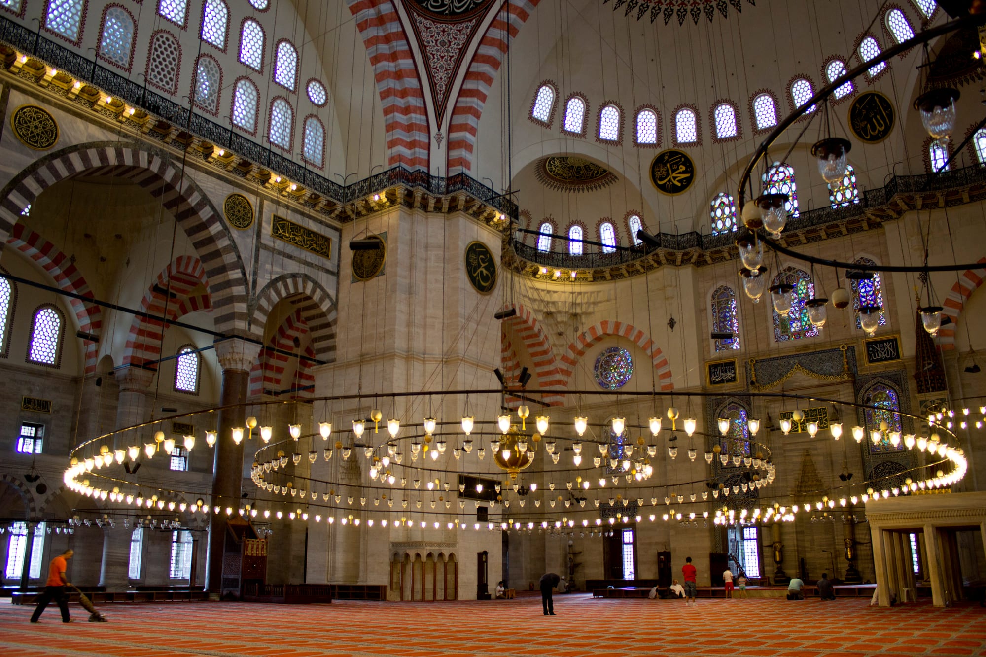 The vast carpeted prayer hall of an Ottoman mosque, with red and white striped arches, rows of stained-glass windows, calligraphy roundels and a huge low ring of hanging lamps, with a few small figures on the carpet below