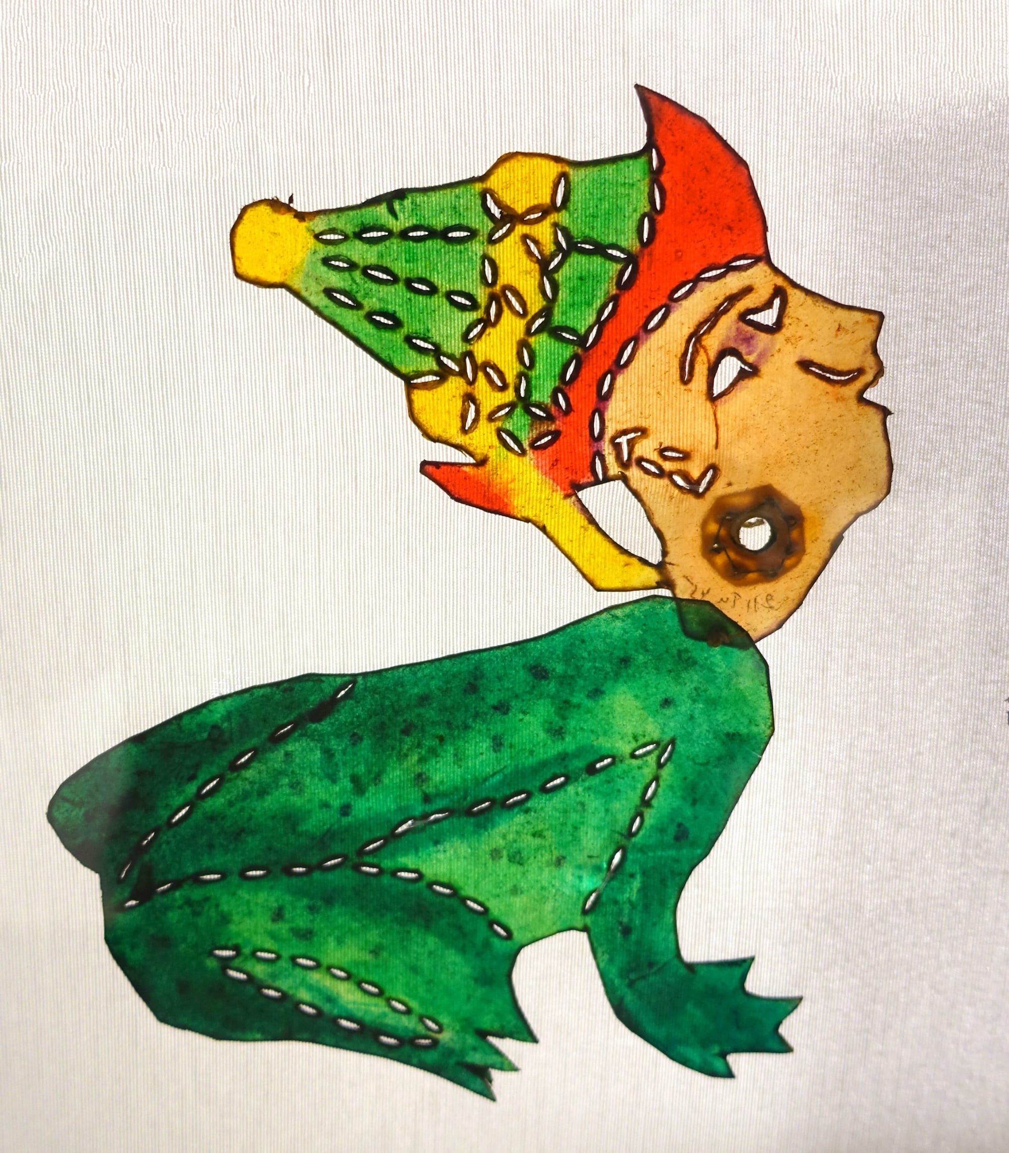 A translucent shadow puppet figure painted in green, orange and yellow, showing a character with a pointed cap whose body has been turned into a frog, lit from behind against a white screen