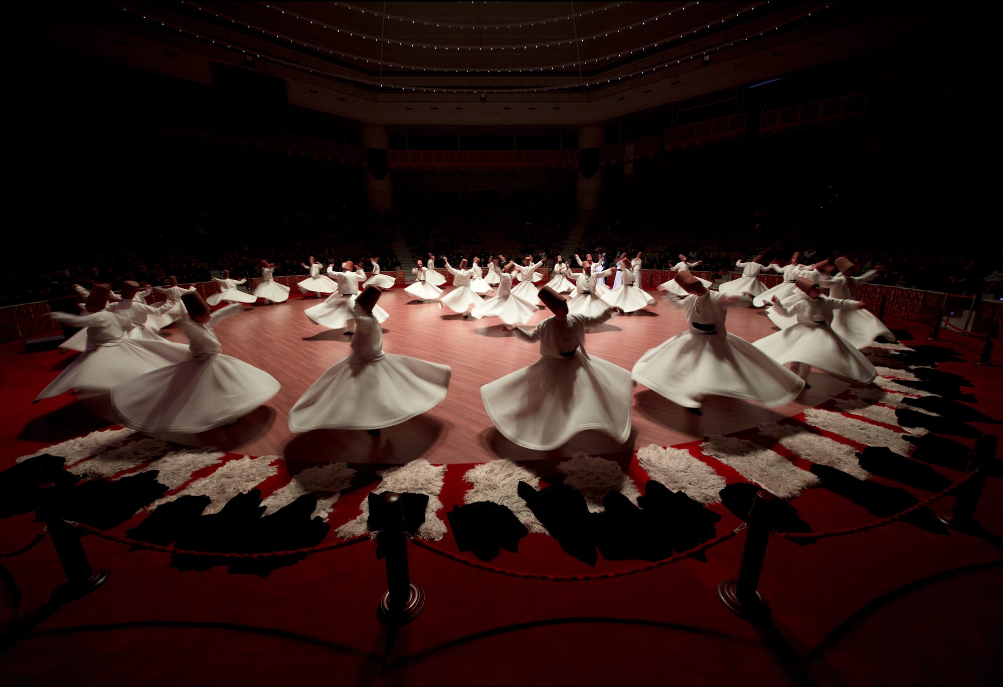 Dozens of dervishes in white robes and tall brown felt hats whirl across a round wooden floor edged with sheepskins, their skirts flared out around them, in a darkened hall