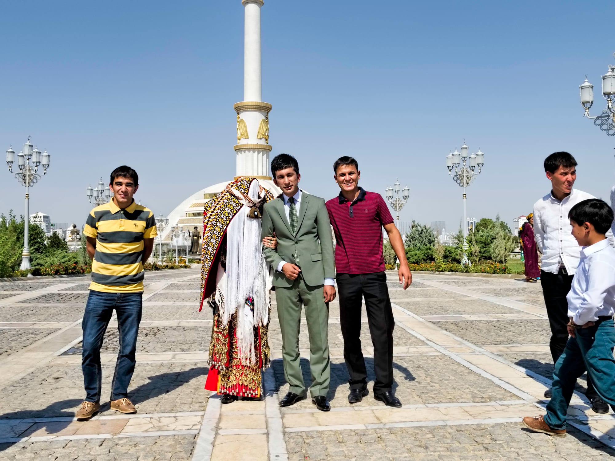 A group of young men and boys standing and smiling near a tall white column monument in an Ashgabat plaza