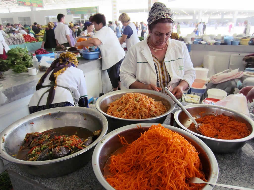 Vendors in headscarves selling large bowls of pickled vegetables and salads at a busy covered market in Ashgabat