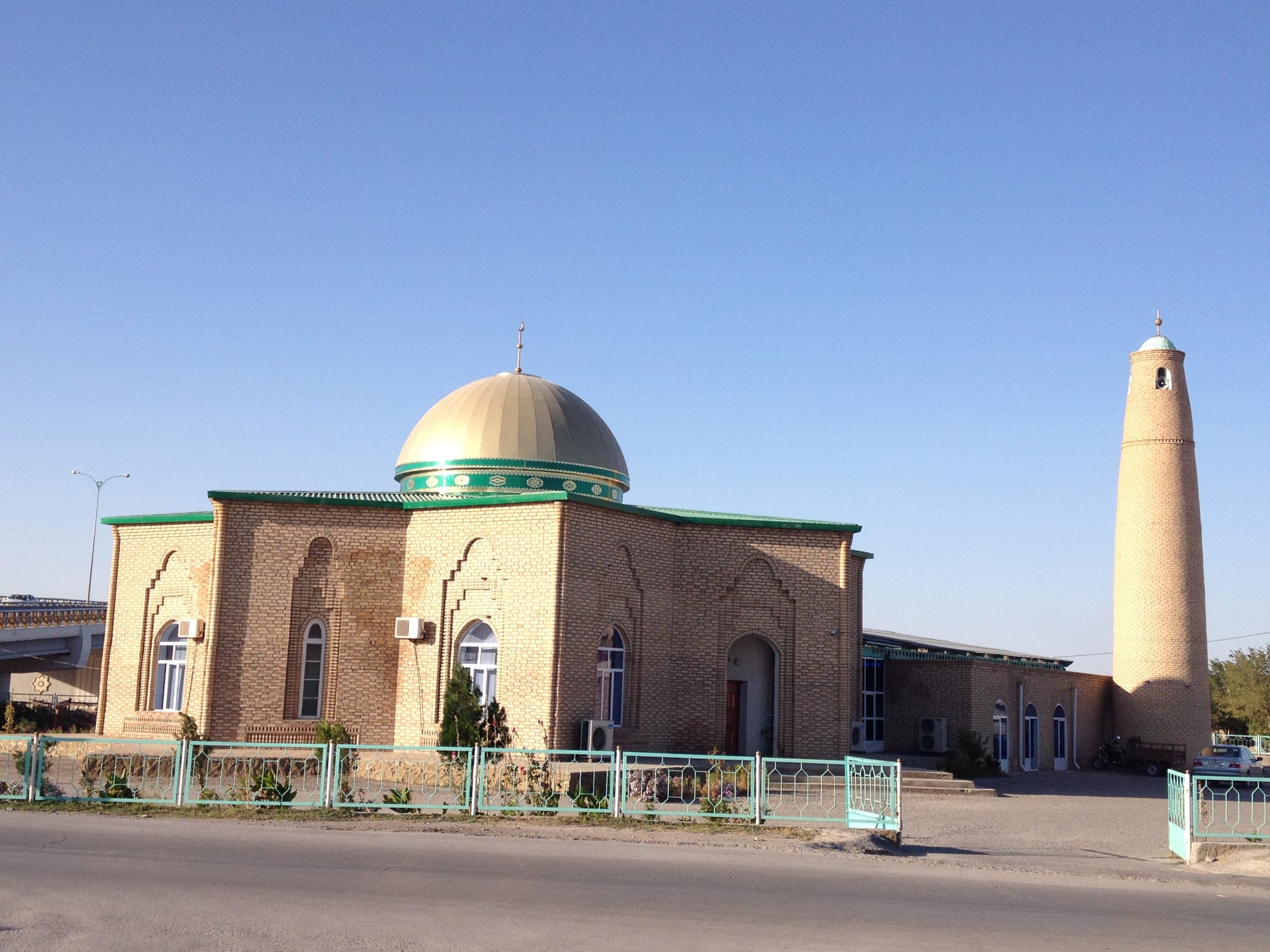 A brick mosque with a golden dome and green-trimmed roof behind a green railing, with a tall round brick minaret standing to the right under a clear blue sky