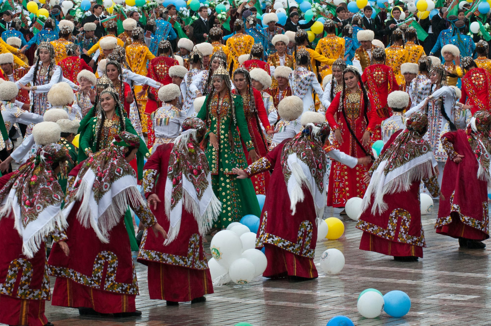 Women in long embroidered red and green dresses and fringed shawls dance in a paved square among coloured balloons, with rows of performers in white sheepskin hats and bright robes behind them