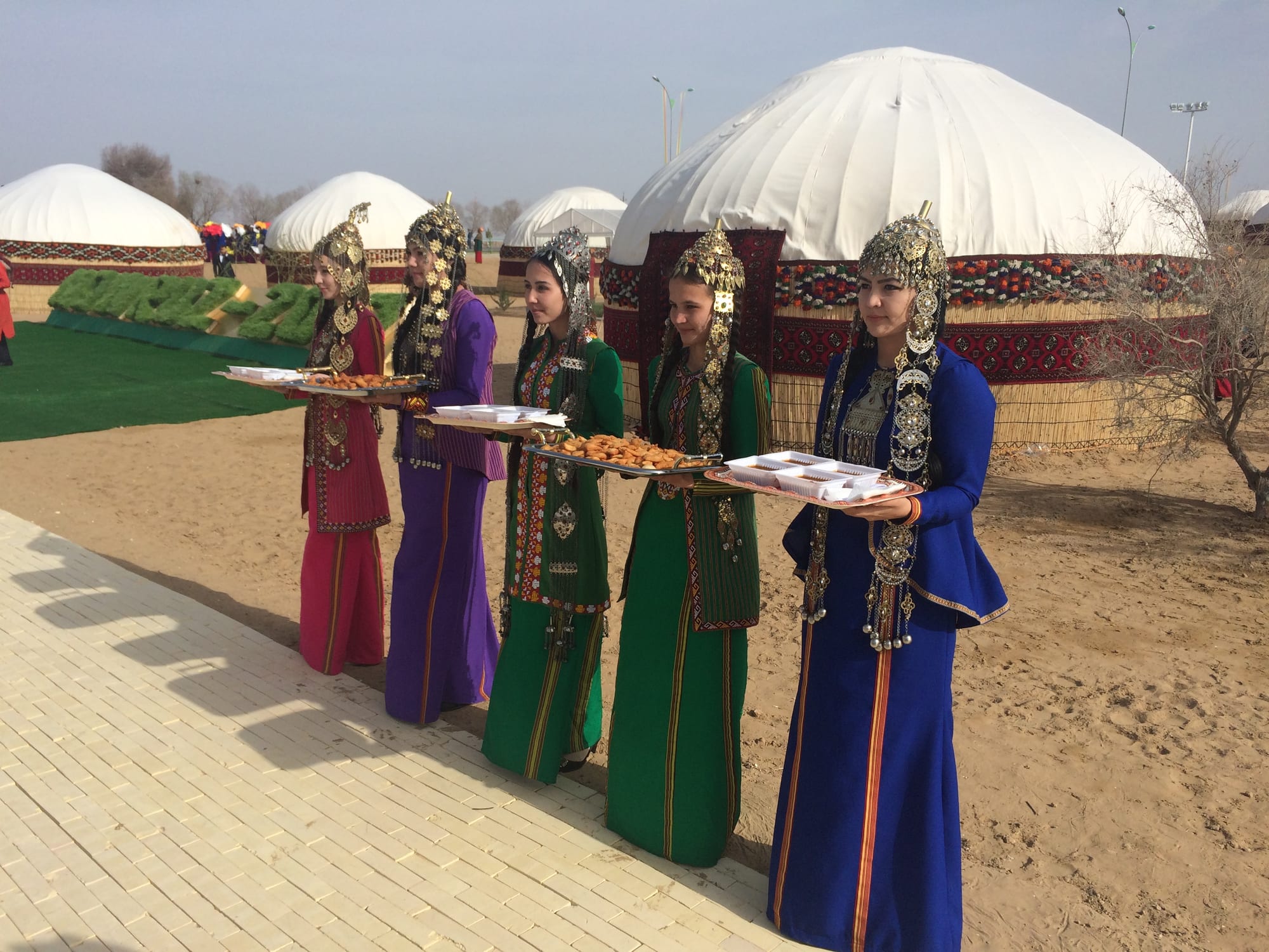Five young women in long dresses of red, purple, green and blue, with tall silver headdresses and jewellery, stand in a line holding trays of food in front of white yurts decorated with red bands